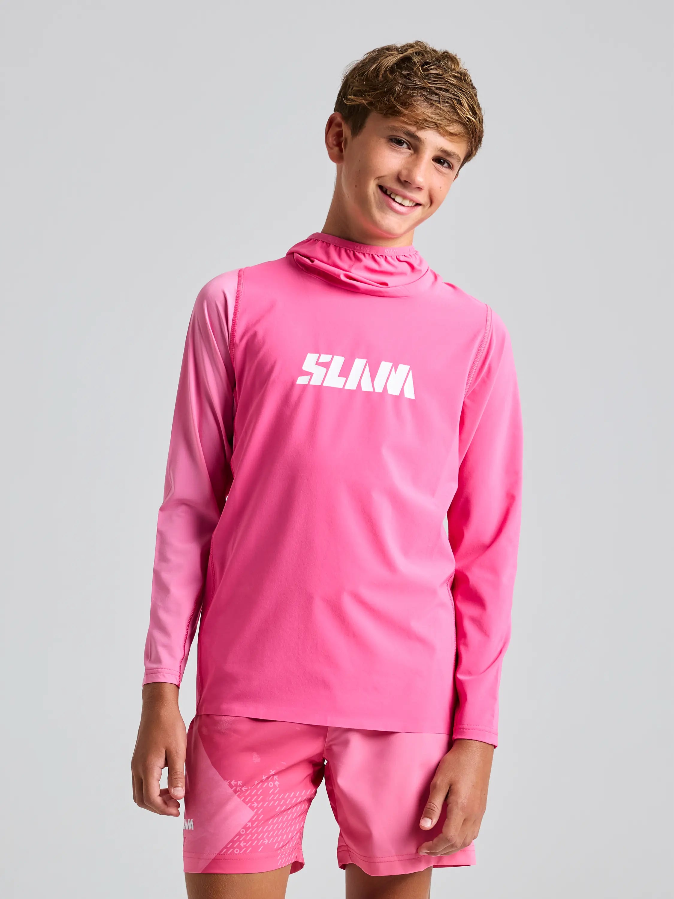 They're wearing a vibrant SLAM OD JR TECH HOODED T-SHIRT, made from recycled stretch fabric, in bright pink with "SLAVA" boldly printed on it and matching shorts. Standing against a plain gray background, they smile slightly, embodying vibrant sustainability.