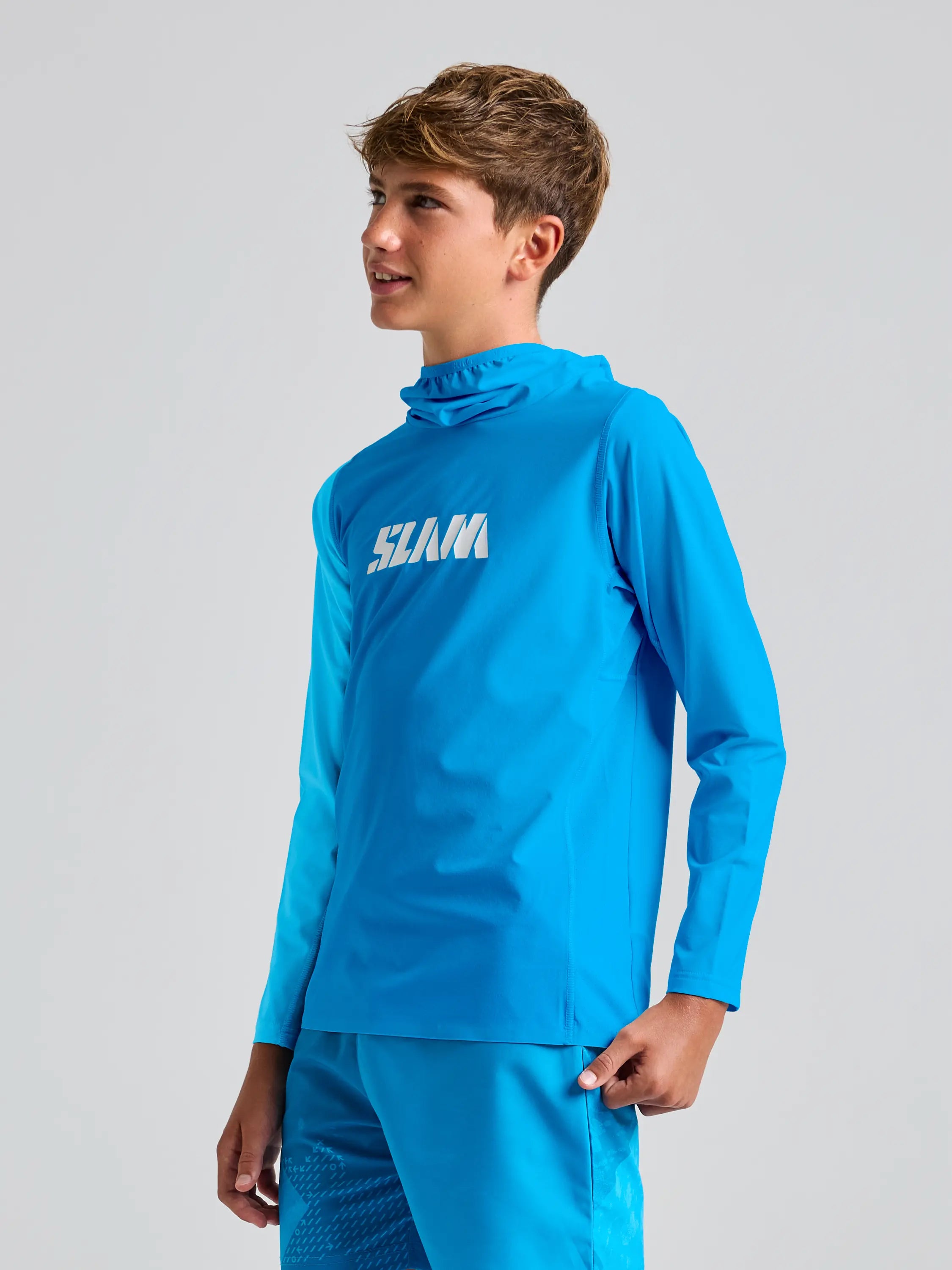 A young person in a blue OD JR TECH HOODED T-SHIRT by SLAM, made from recycled stretch fabric, gazes sideways. The top is coupled with matching blue shorts featuring 50+ UPF protection, all set against a plain, light-colored backdrop.