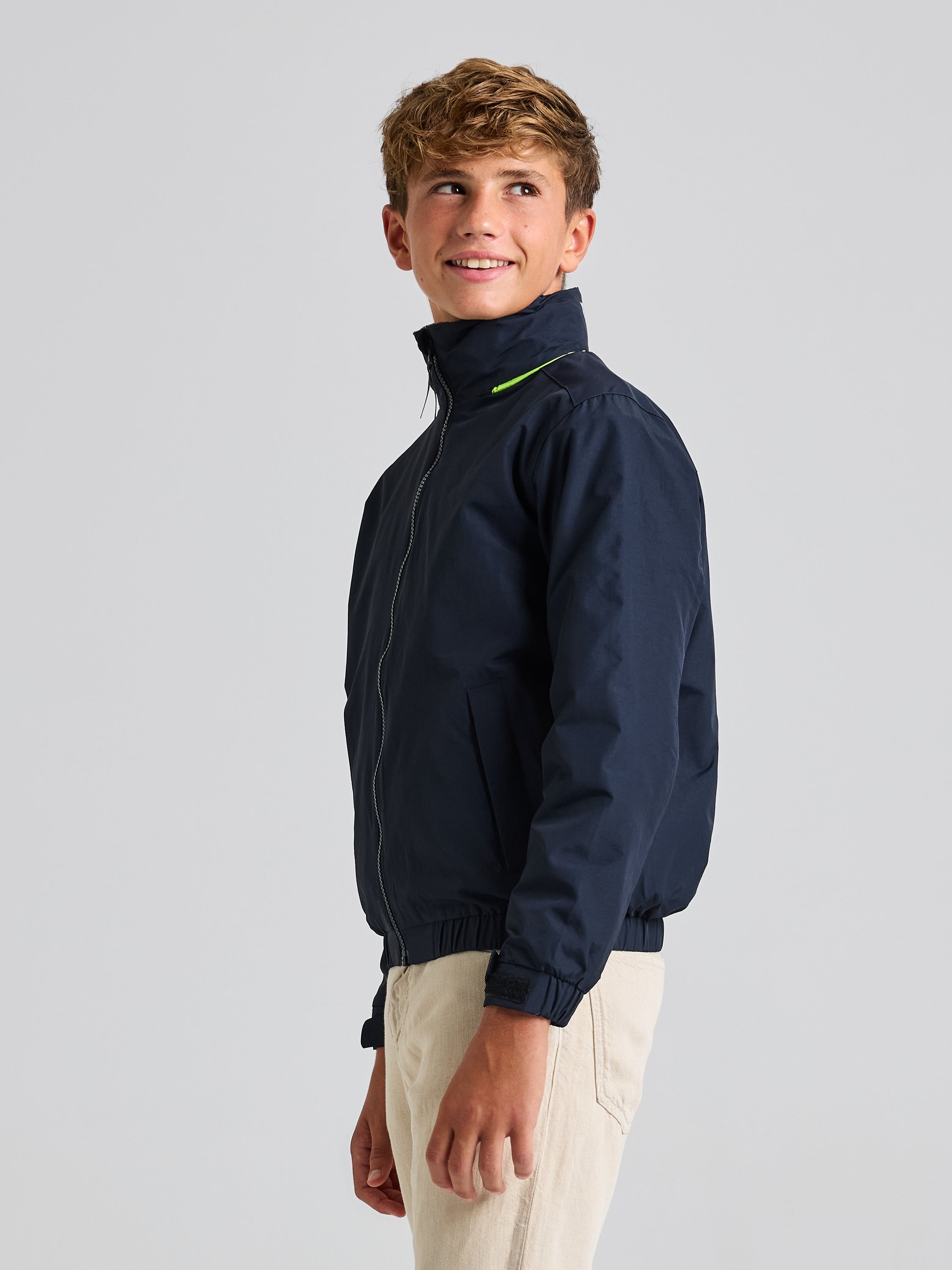 A young person with short, tousled hair is wearing the SLAM JUNIOR SAILING WARM JACKET in navy, which is water-resistant and features a sleek zipper. They are smiling while looking over their shoulder, standing against a plain background. The jacket is made from Taslon 228T nylon and boasts a distinctive neon detail near the collar.