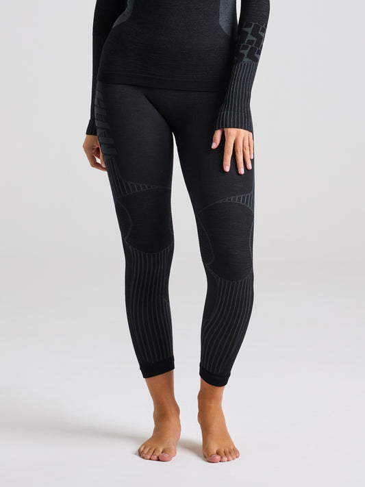 A person is standing barefoot, dressed in SLAM's PRO SEAMLESS MERINO TIGHT, featuring black ink thermal leggings and a matching long-sleeve top made from Merino-blend yarn. This comfortable undergarment showcases subtle patterns with ribbed details on the knees and elbows against a plain light gray background.