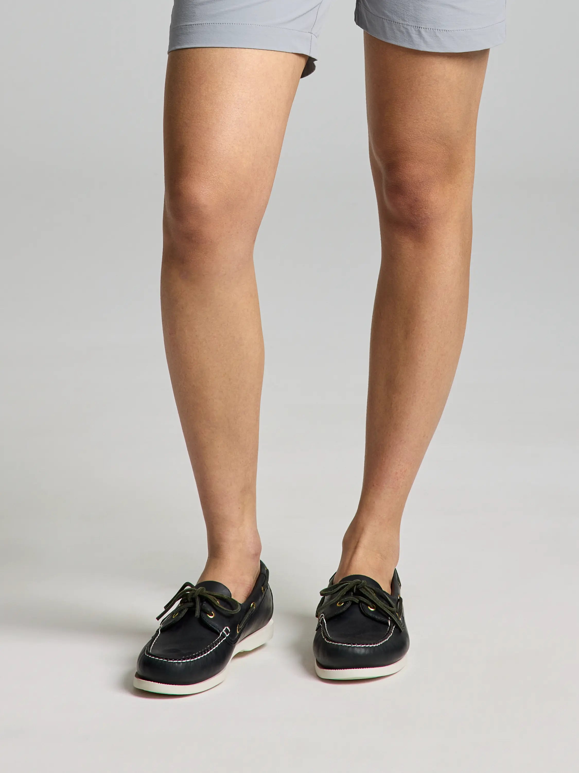 A person's legs are shown wearing light gray shorts and stylish slip-on boat shoes designed by SLAM. These BOAT SHOES come in DARK NAVY with white soles and tan laces, set against a plain, neutral background.