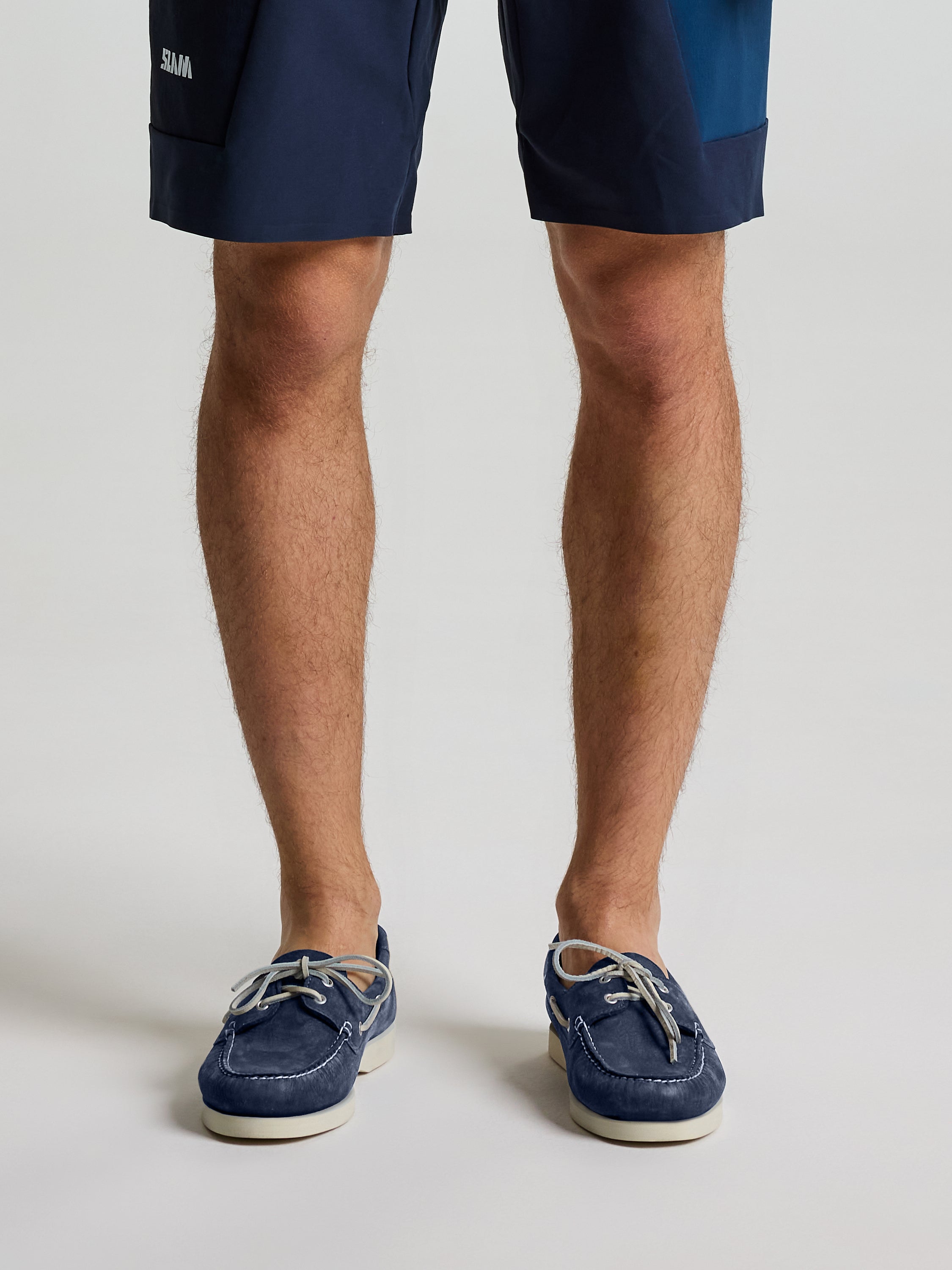 A person models the lower half of their legs against a plain light background, wearing SLAM NABUCK BOAT SHOES in DARK NAVY paired with navy blue shorts. The shoes feature non-slip soles.