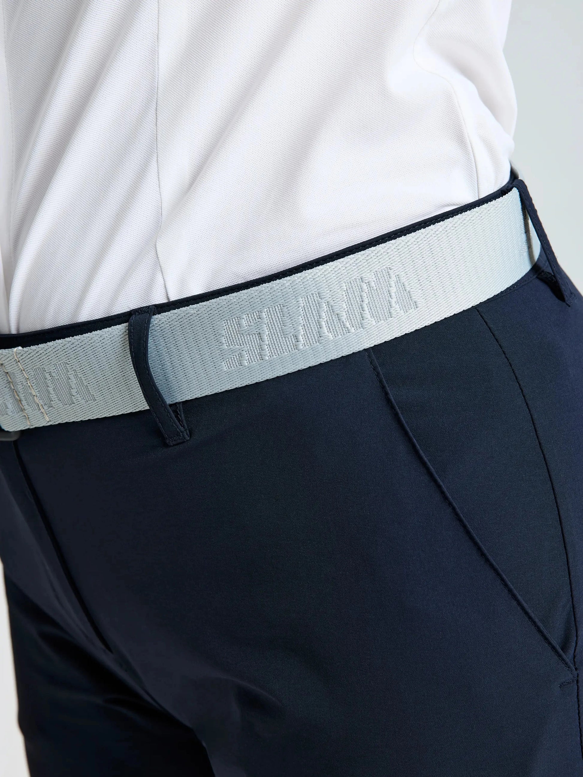 A person is wearing a white shirt tucked into dark navy technical trousers, accessorized with the SLAM LOGO BELT 2.0 in light gray. The focus is on the waist area, highlighting the adjustable belt and a partially visible woven SLAM label.
