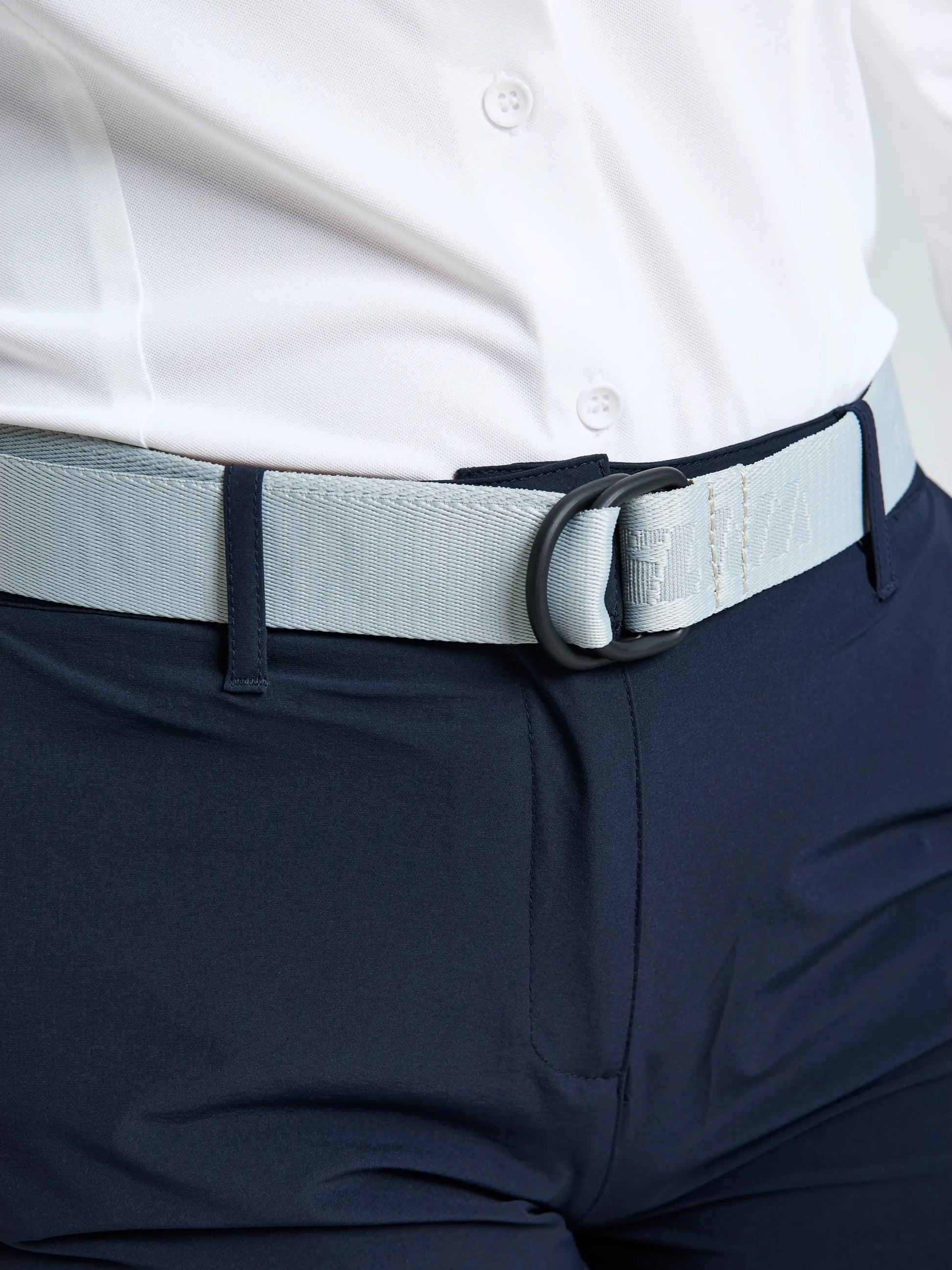 A person wearing a white button-up shirt, navy technical trousers, and the SLAM LOGO BELT 2.0—a light gray adjustable belt with a black D-ring buckle. The image is cropped to focus on the waist and the SLAM LOGO BELT 2.0.