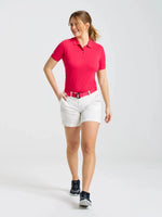A woman wearing a bright pink polo shirt, white shorts with the SLAM LOGO BELT 2.0 in pink, and navy sneakers walks on a plain white background, looking to the side and smiling.