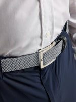 A close-up of a person wearing a white shirt, navy pants, and the SLAM DECK BELT in ALUMINIUM—a light gray woven elastic belt with a rectangular metal buckle—creating a classic nautical look.