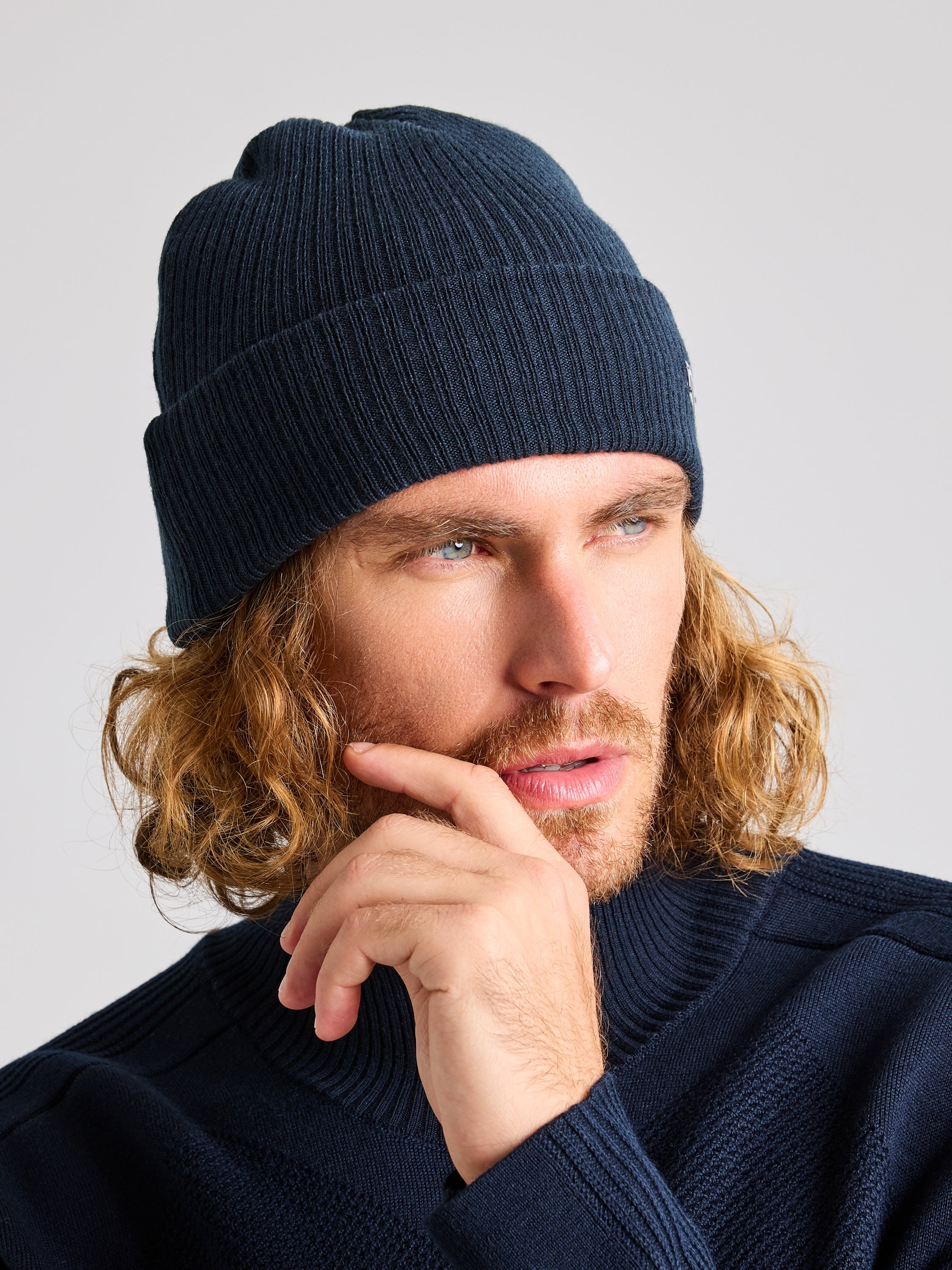 A person with long curly hair is wearing a dark blue unisex winter CABLE BEANIE by SLAM, along with a matching sweater crafted from recycled yarn. They are gazing thoughtfully to the side, hand near chin, against a plain background.