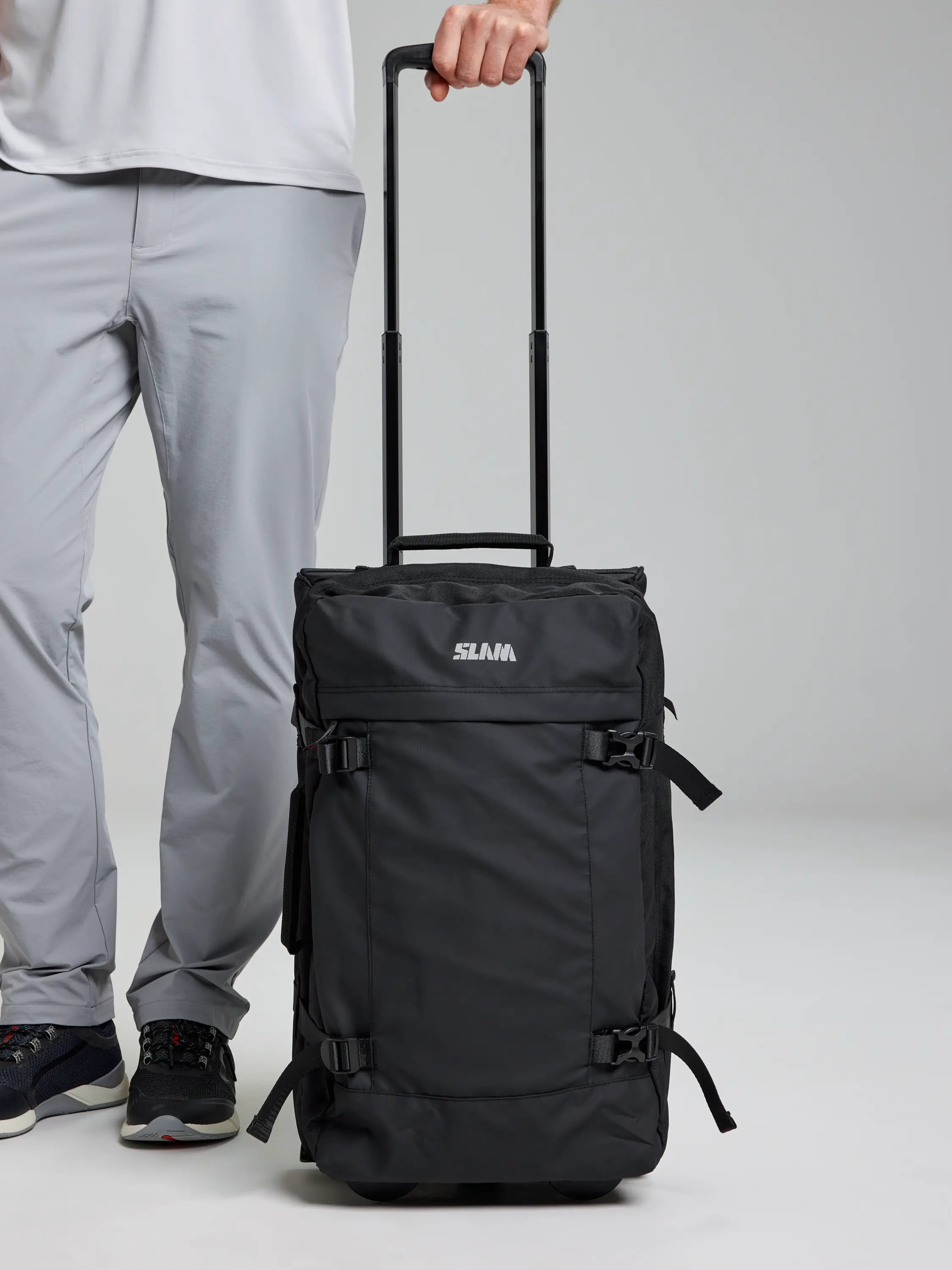 A person wearing light gray pants and black sneakers holds the extended handle of a sleek unisex CABIN TROLLEY with the brand "SLAM" printed on it. Perfect for a short trip, this sturdy suitcase in BLACK INK features multiple buckles, ready for any weekend on the boat.