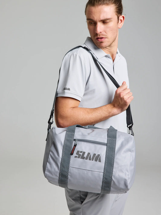 A person with long hair is wearing a light gray polo shirt and pants, accessorizing with the WR Duffle Bag S by SLAM. This aluminum-colored, water-repellent bag features dark gray handles and a shoulder strap, all set against a plain, light background.