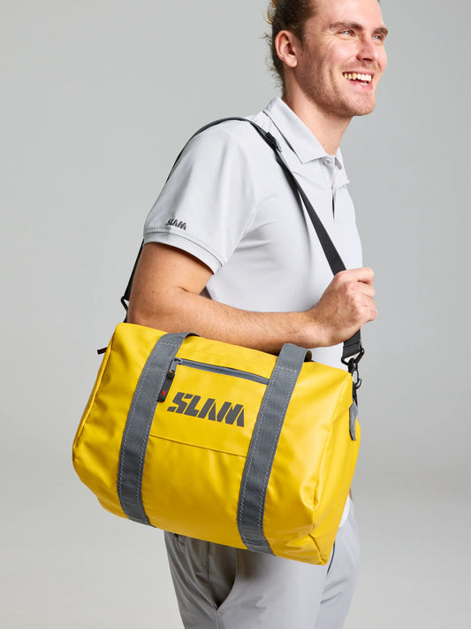 Dressed in a light grey polo shirt and pants, a person carries the WR Duffle Bag S by SLAM in an ocher hue. The water-repellent bag enhances their subtle nautical style as they smile, looking to the side.