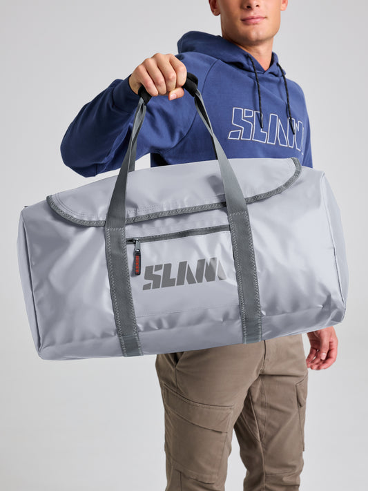 A person in a blue hoodie and khaki pants holds up the SLAM WR DUFFLE BAG L in ALUMINIUM, featuring dark gray straps and a front zipper pocket. The light gray waterproof bag is branded with “SLAM” and built for any weather.
