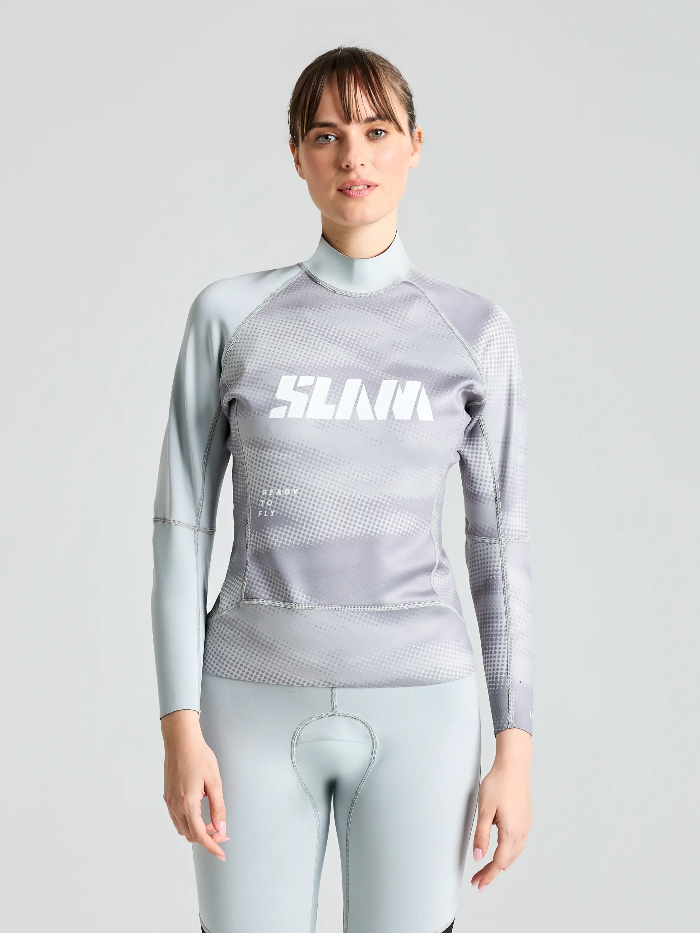 A person in a gray FOIL TOP 1.5 by SLAM poses against a plain background. The long-sleeve wetsuit, featuring targeted compression, showcases a geometric pattern and "SLAM" boldly printed on the chest. The individual sports short hair and gazes confidently at the camera.