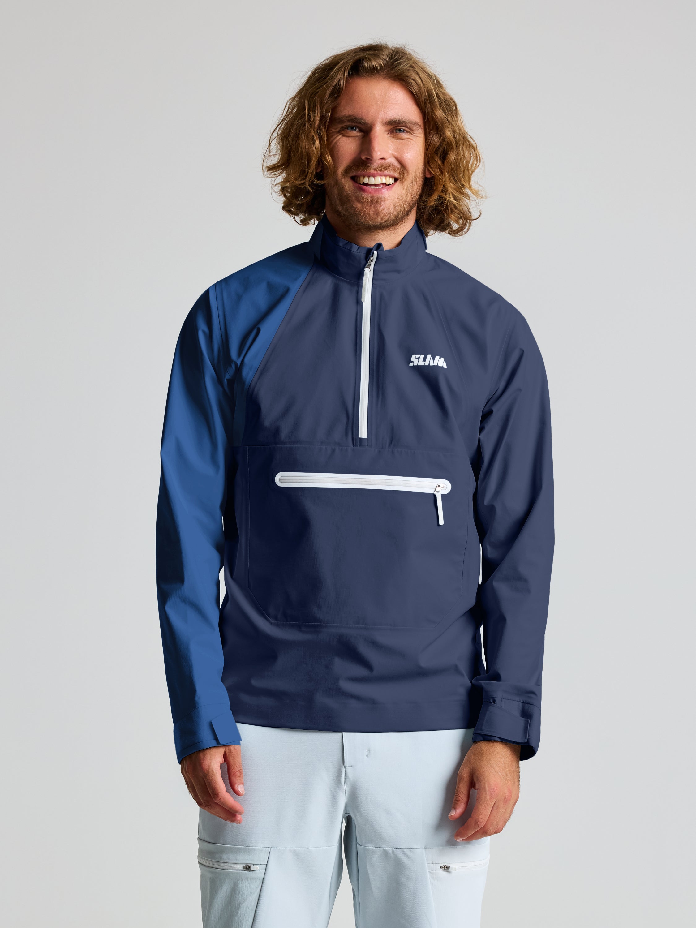 A smiling man with wavy light brown hair and a beard wears a SLAM FOIL SPRAY TOP pullover jacket in DARK NAVY with waterproofing and a white zipper pocket, paired with white pants, standing against a plain background.