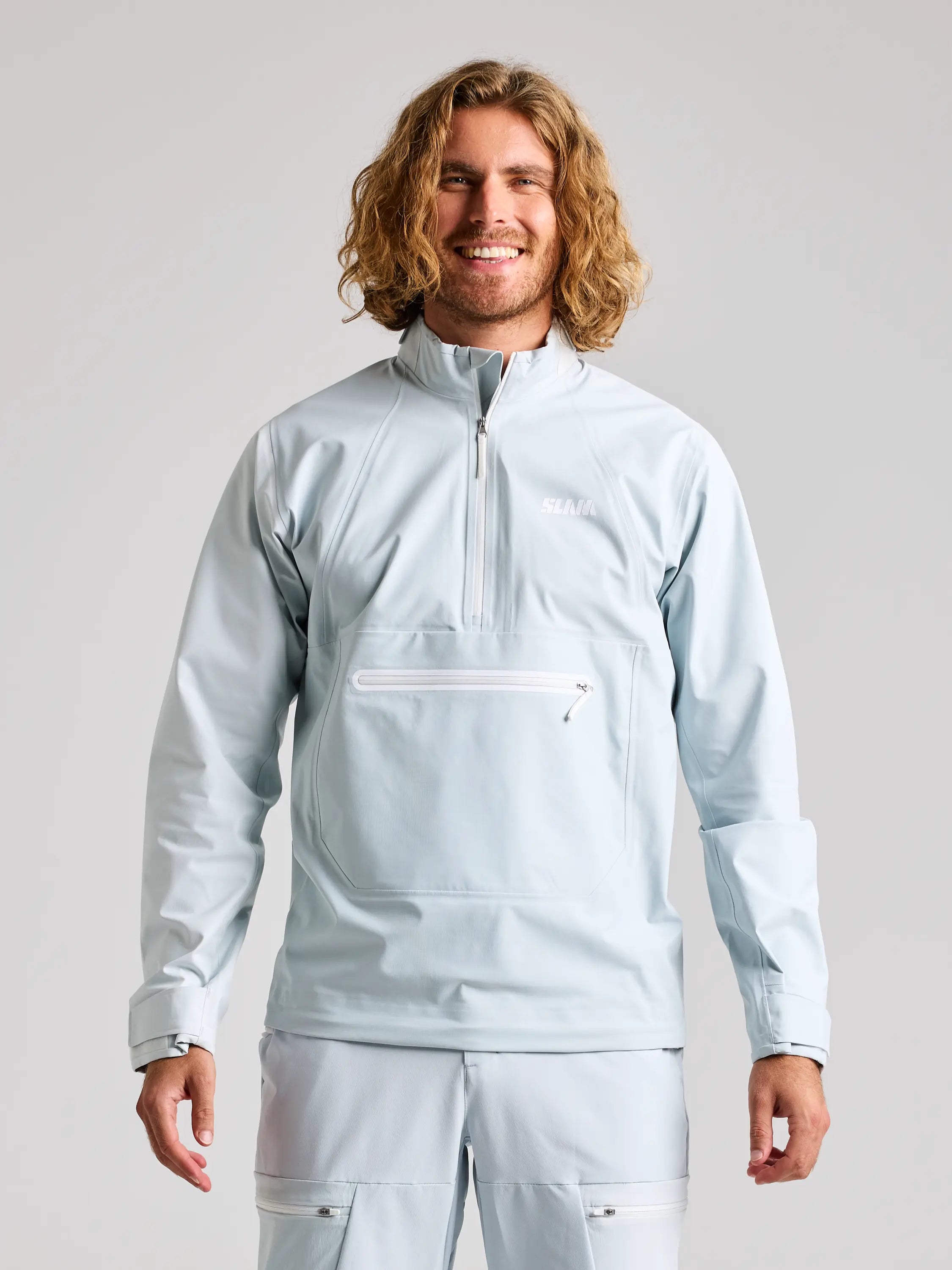 A person with long wavy hair smiles while wearing the SLAM FOIL SPRAY TOP in CLIFF GREY, showcasing a zippered pocket and high breathability for comfort. The background is plain and light-colored.