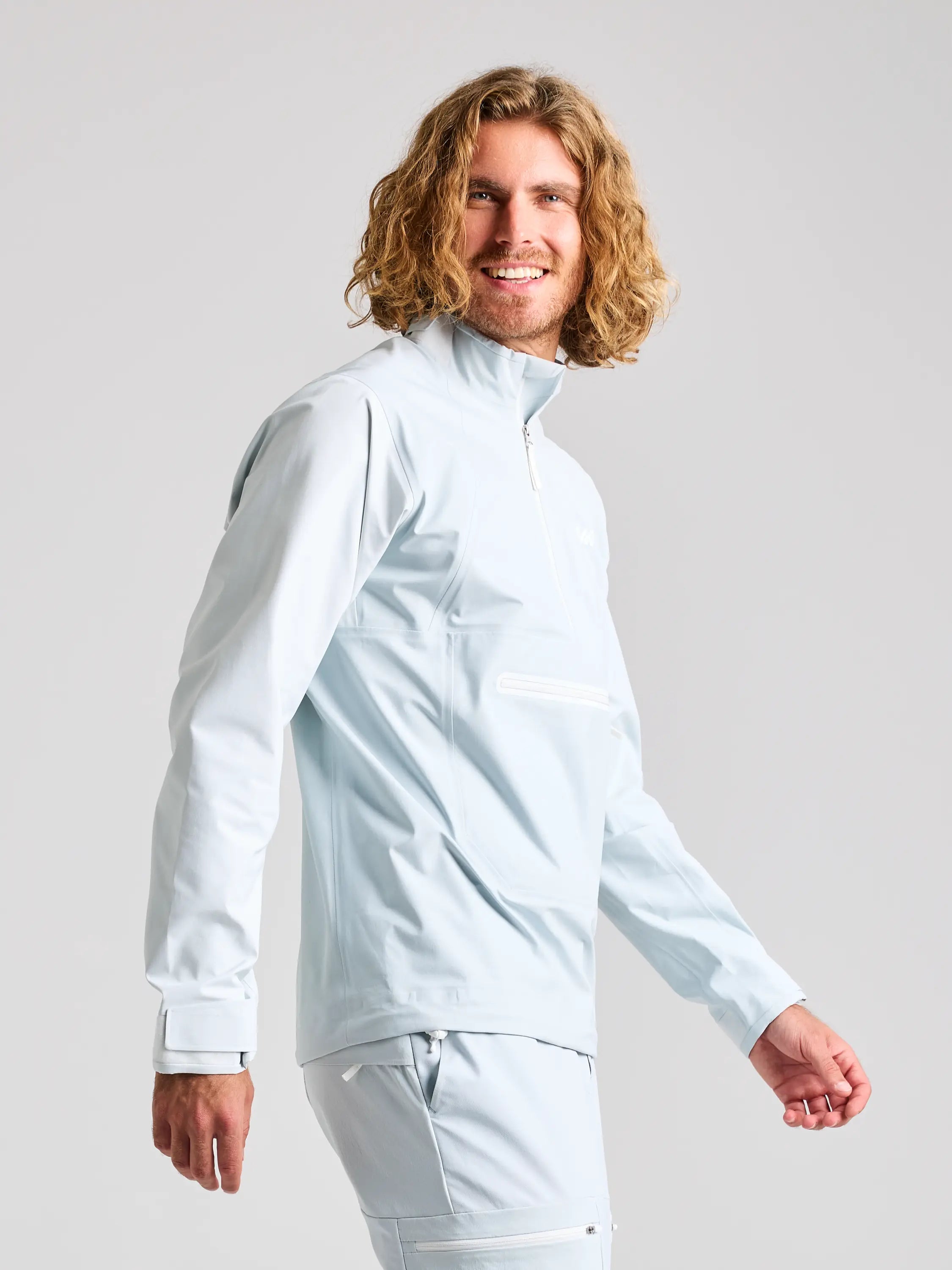 A person with long curly hair smiles while wearing the SLAM FOIL SPRAY TOP, a light blue zip-up jacket praised for its high breathability. They are also in matching pants, standing against a plain gray background, looking slightly to the left.