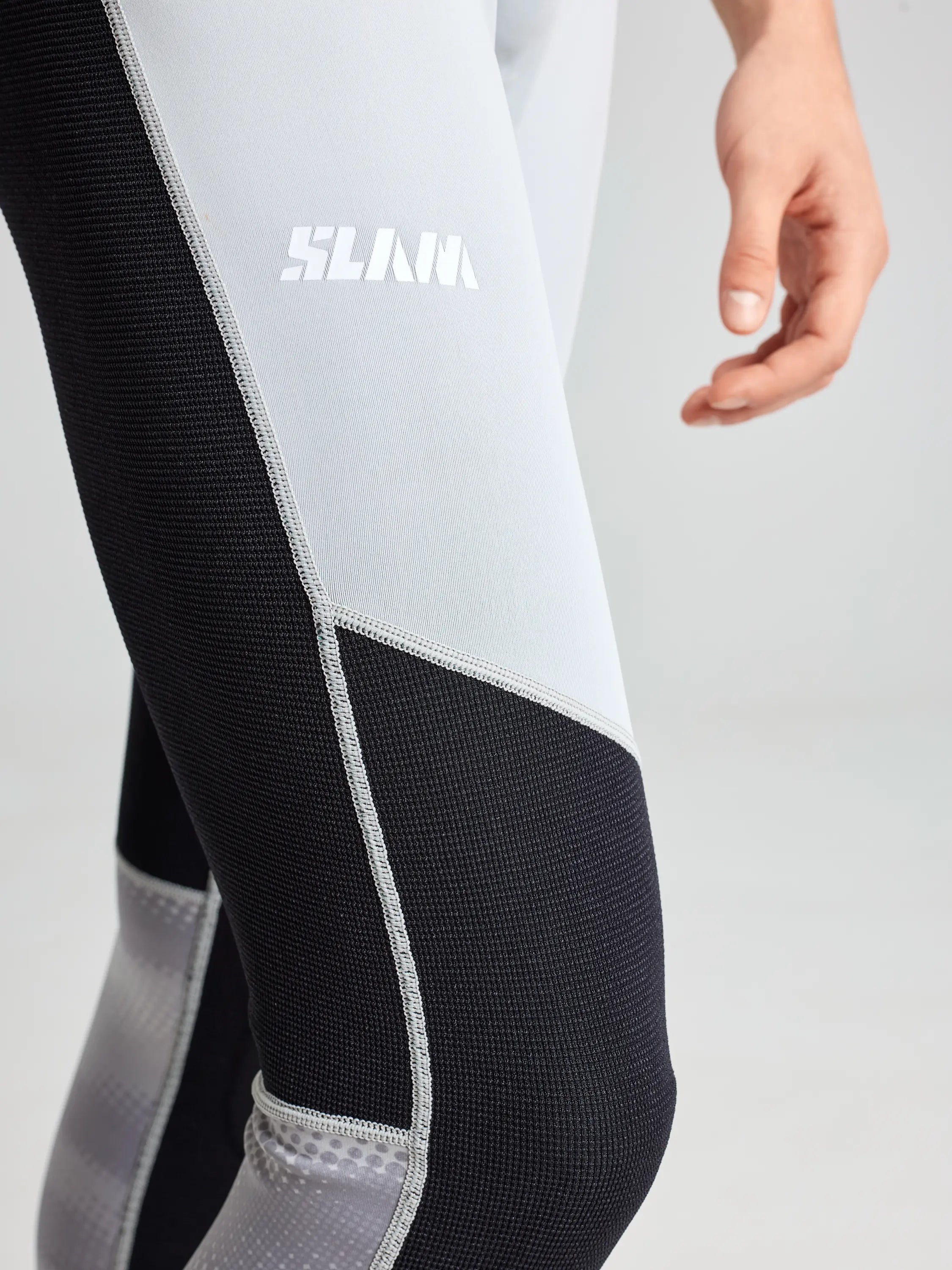 Close-up of a person in SLAM's FOIL LONG JOHN 1.5 black and light gray athletic leggings, ideal for foiling or extreme water sports. A white "SLAM" logo stands out on the side against the plain light gray background; their arm is slightly bent, fingers relaxed.