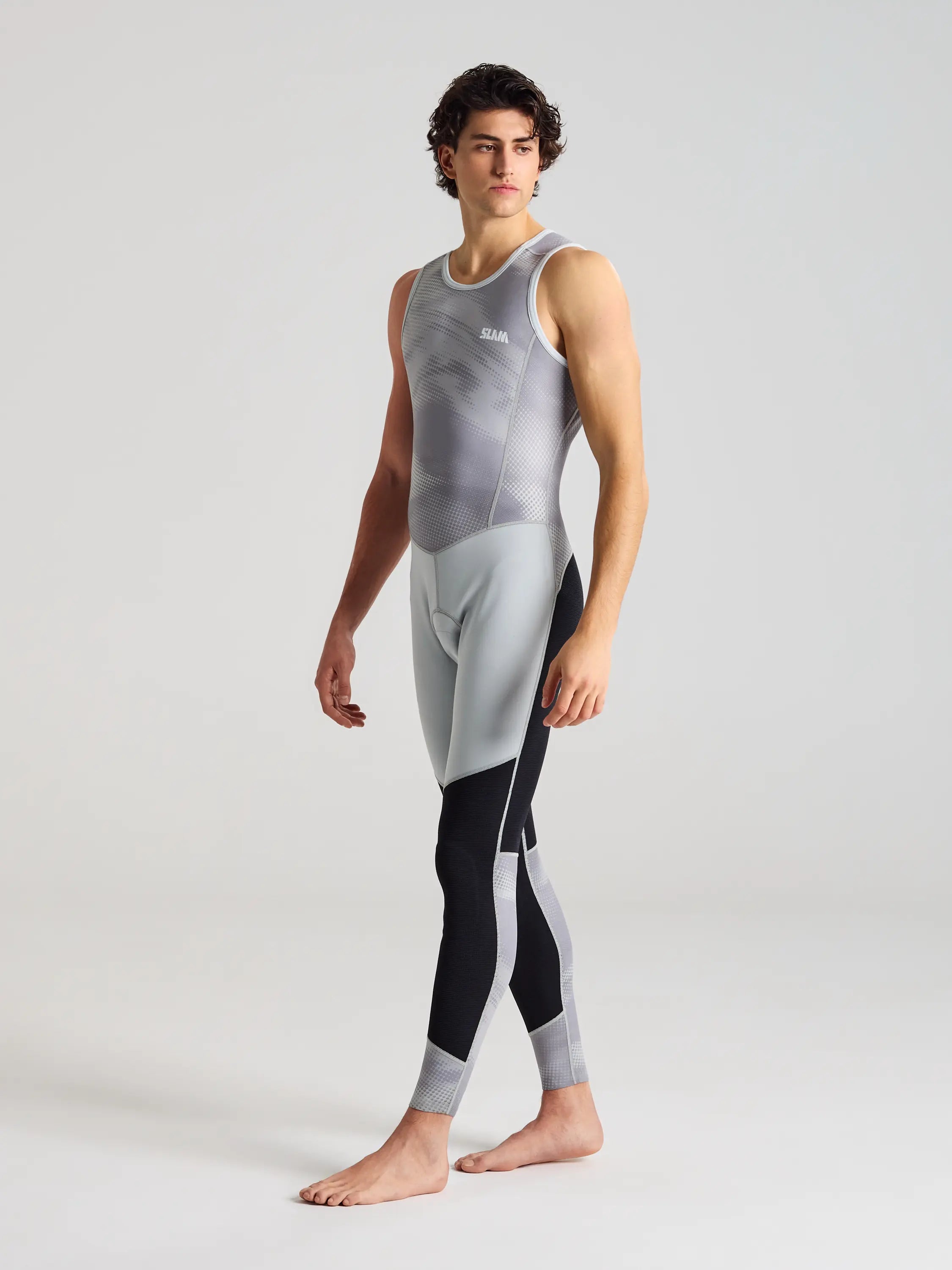 A person stands barefoot against a plain background, wearing SLAM's FOIL LONG JOHN 1.5 in gray and black neoprene. With a neutral expression and gaze to the side, they evoke athletes ready for extreme water sports.