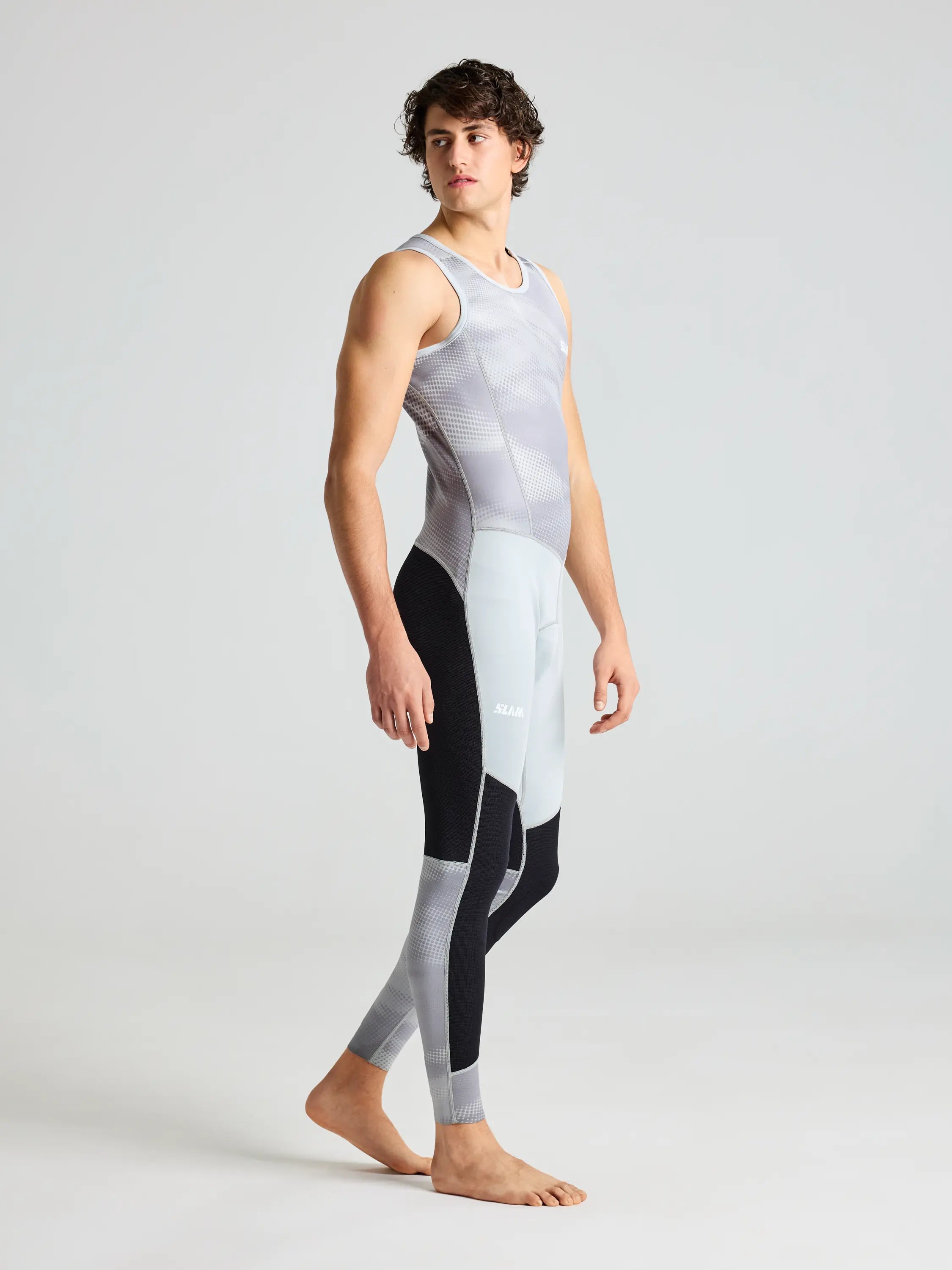 A person stands barefoot wearing the sleeveless, form-fitting SLAM FOIL LONG JOHN 1.5 bodysuit with a gray and black pattern, resembling neoprene long johns for extreme water sports. The background is plain and light-colored.