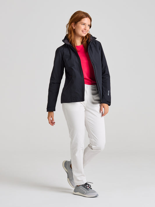 A person stands against a plain background wearing a SLAM CREW WOMAN WARM HOODED JACKET, a pink shirt, white pants, and gray sneakers. The jacket's waterproof material adds practicality to the ensemble. They have a relaxed stance with one hand in a pocket.