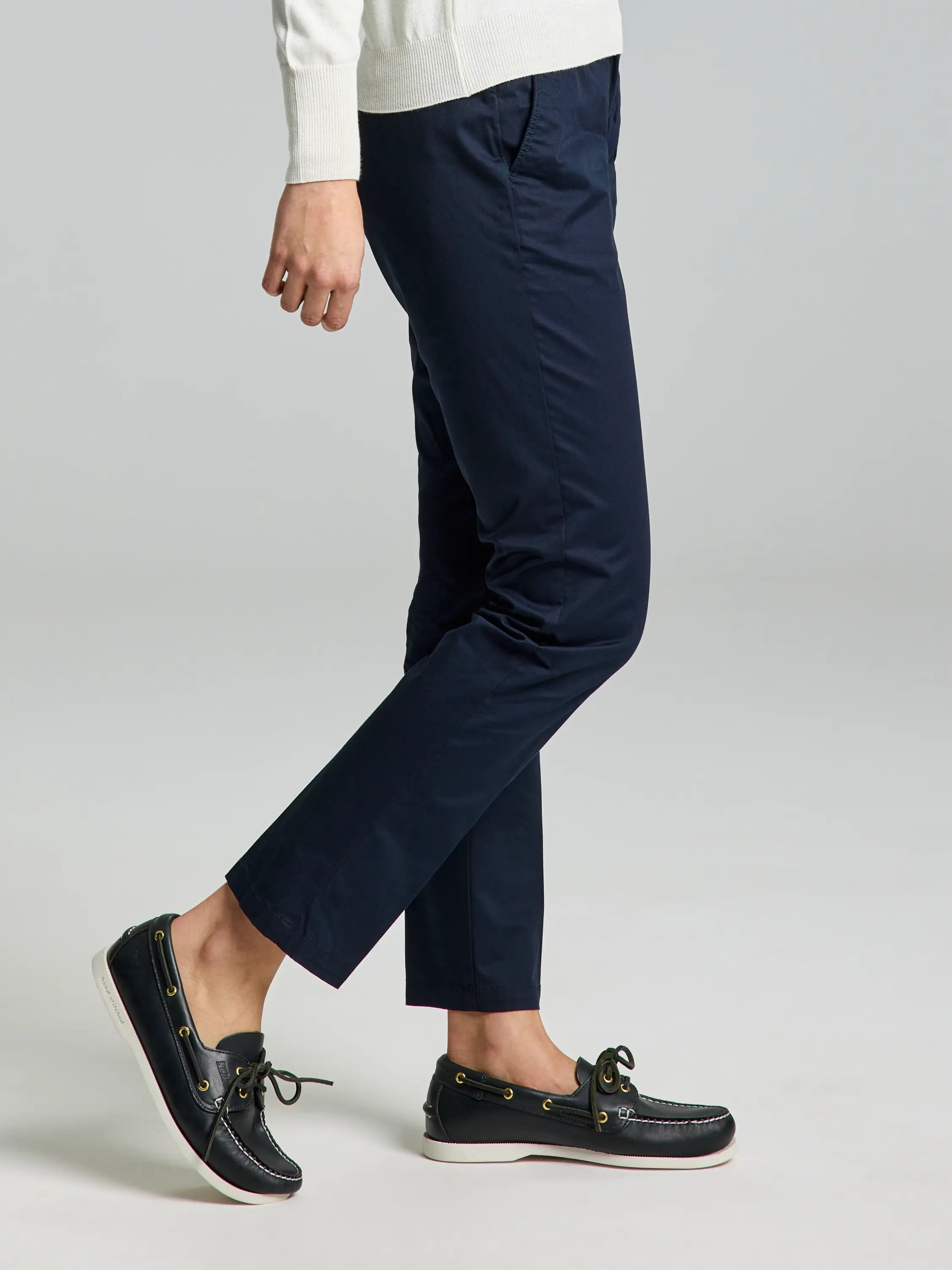 A person wearing a white sweater, DECK WOMAN LIGHT CHINO PANT in navy blue by SLAM, and black boat shoes stands against a plain gray background. The image captures a side view from the shoulders down, showcasing the essence of formal summer wear.