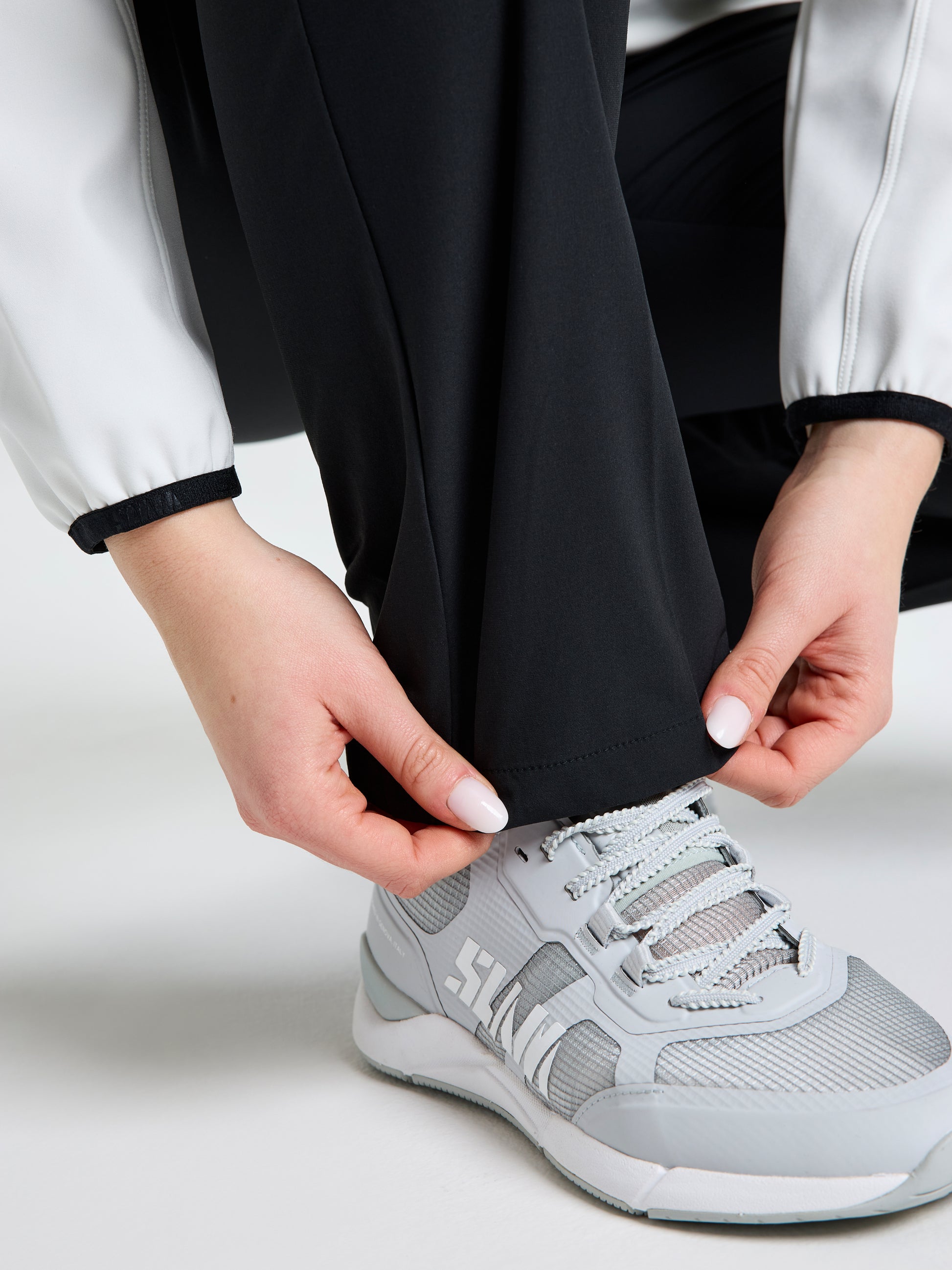 A person with white nail polish adjusts the cuff of SLAM's lightweight WS TECH CHINO PANT in black, paired with a white and gray athletic shoe. The plain white background highlights the sleek style of these women's trousers.