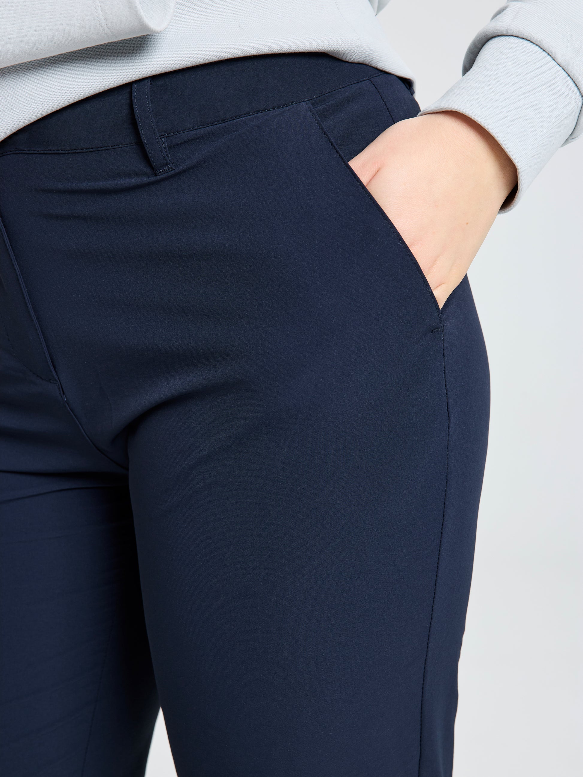 A person models the SLAM WS TECH CHINO PANT in navy blue, standing with one hand in their pocket. The cropped image focuses on the comfort and style of these lightweight women's trousers.