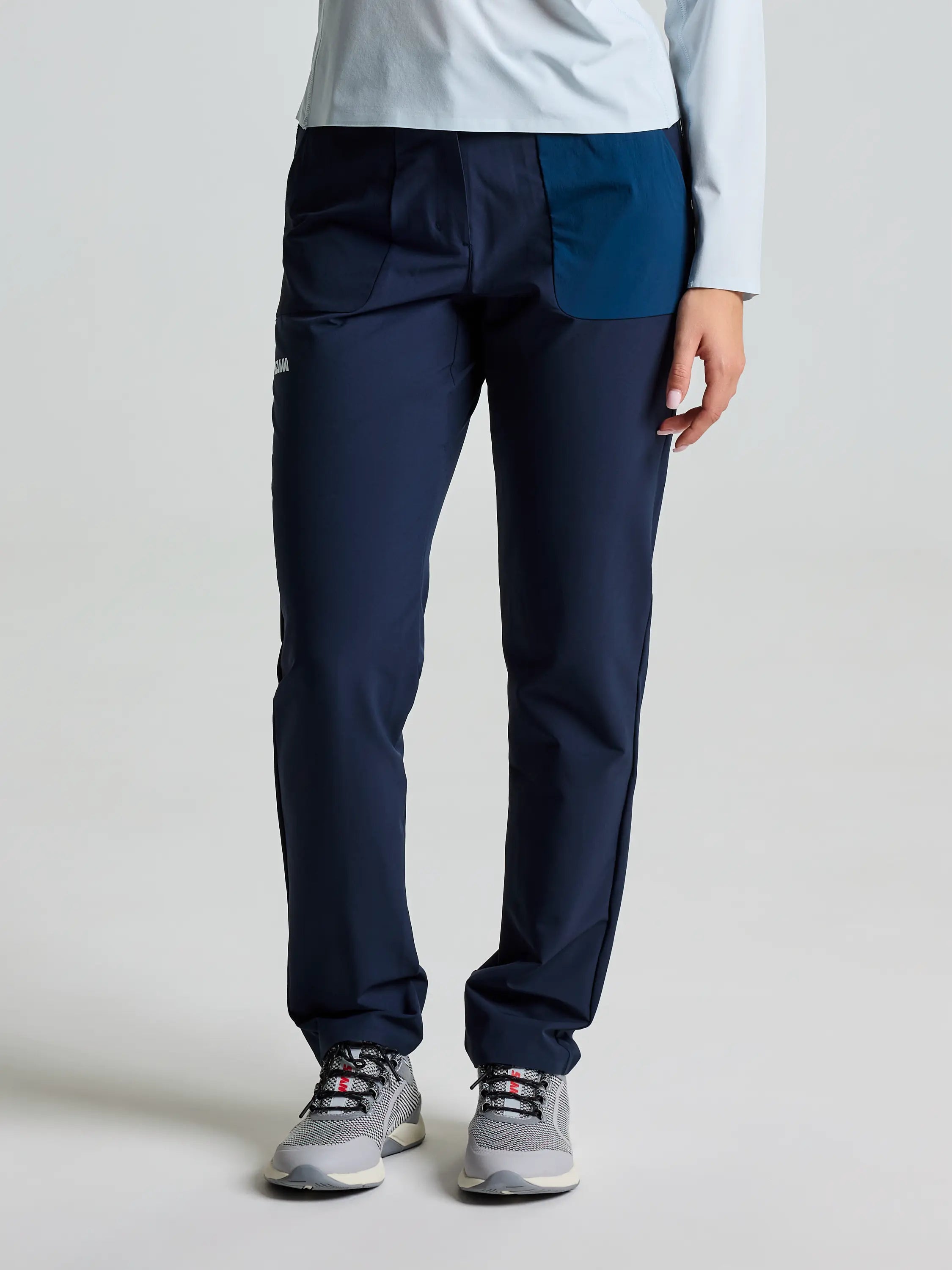 Someone is dressed in SLAM's OD WS CHINO PANT in DARK NAVY with a light blue long-sleeve shirt, complementing gray sneakers with white soles. A light gray backdrop highlights just the upper body and legs.