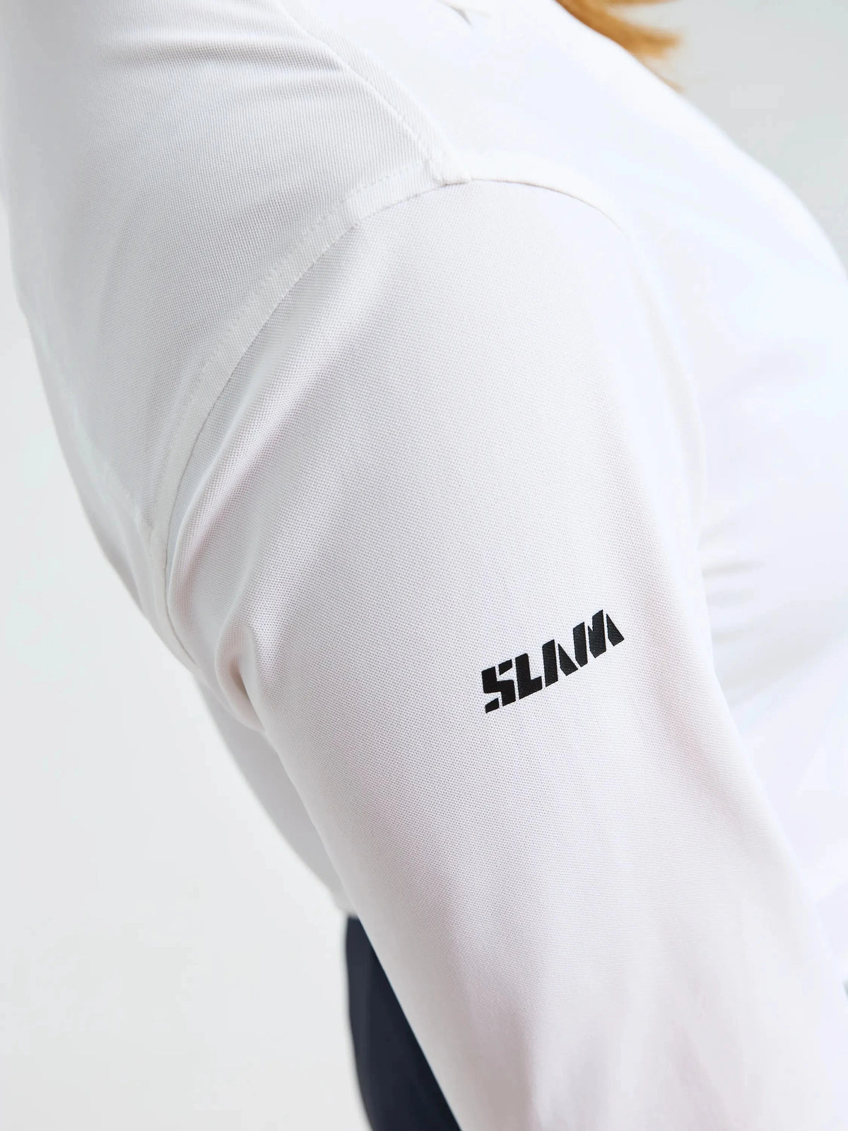 A close-up of a person wearing the SLAM WS TECH PIQUE SHIRT LS in white. The brand name “SLAM” is printed in black capital letters on the upper sleeve. Designed for both style and functionality, this technical long-sleeve shirt provides UPF 50+ sun protection. The background is plain and out of focus.