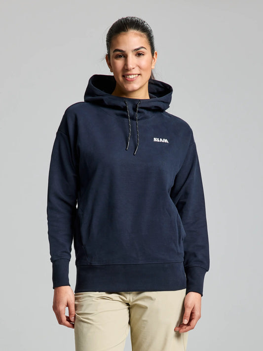 A person wearing a cozy DECK WOMAN HOODIE by SLAM, printed with "SLANG," stands against a plain backdrop. The dark navy hoodie features an adjustable hood and a spacious front pocket. They have long, dark hair pulled back and are paired with beige pants.