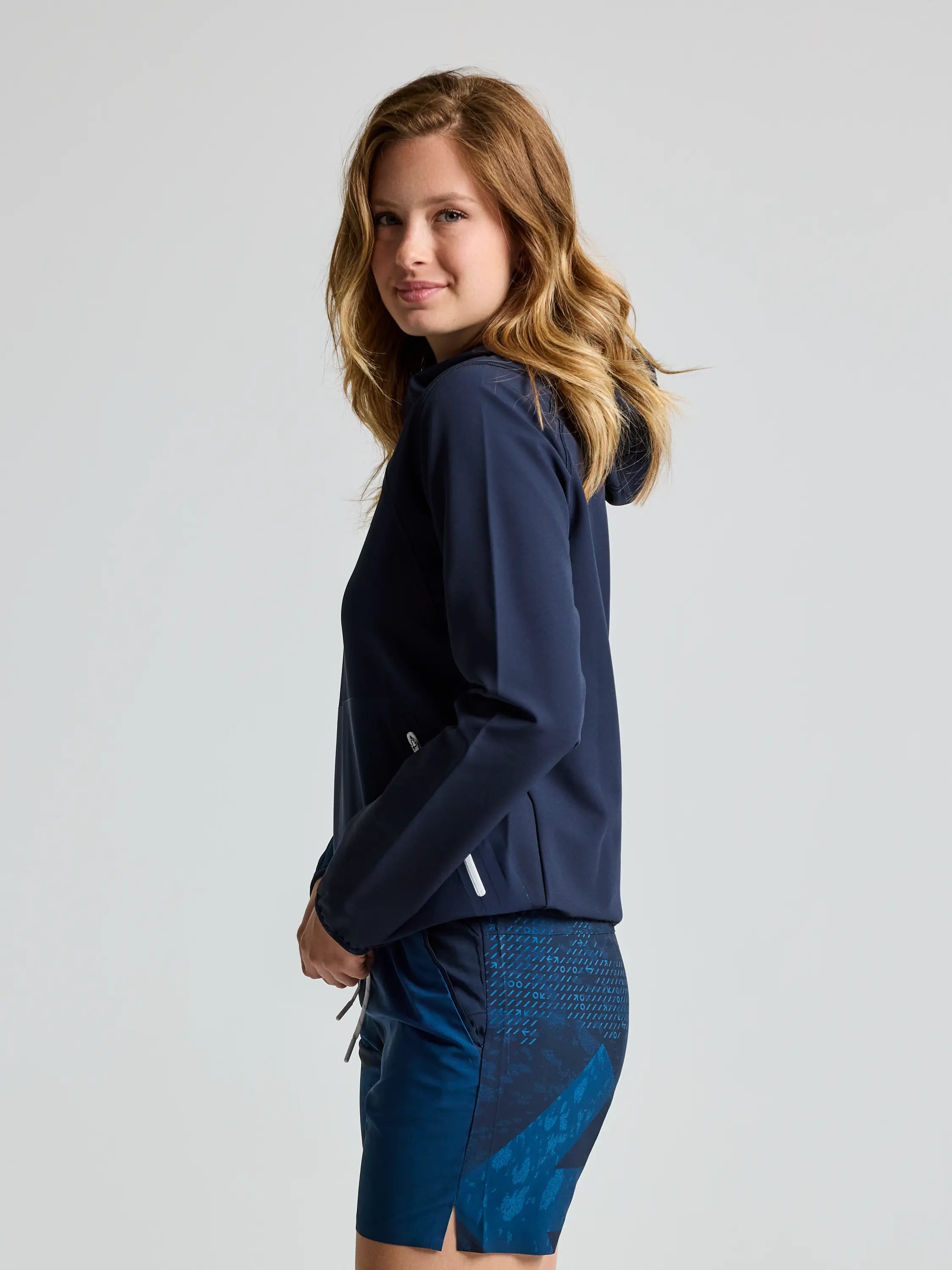 The person with long hair is wearing the SLAM FOIL WS TECH HOODIE, featuring a navy blue color with water-repellent fabric, and paired with blue patterned shorts. They are standing sideways, smiling slightly at the camera against a plain, light-colored background.