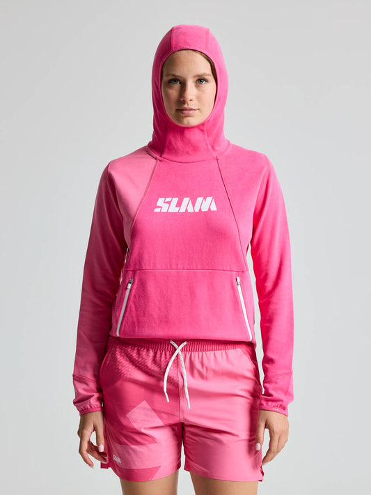 A person sports the GRAPHIC WS HOODIE by SLAM in HYDRANGEA PINK with the hood up, channeling a technical look. Made from bamboo cotton, it has matching shorts, a scuba hood, a large zippered front pocket, and a white logo on the chest against a light-colored background.