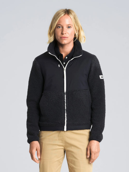 A person with light blonde hair faces forward with a neutral expression, wearing the SLAM ALTURA WS FZ FLEECE—a black zip-up jacket made from warm fleece for added wind protection—paired with tan pants. The background is plain and light gray.