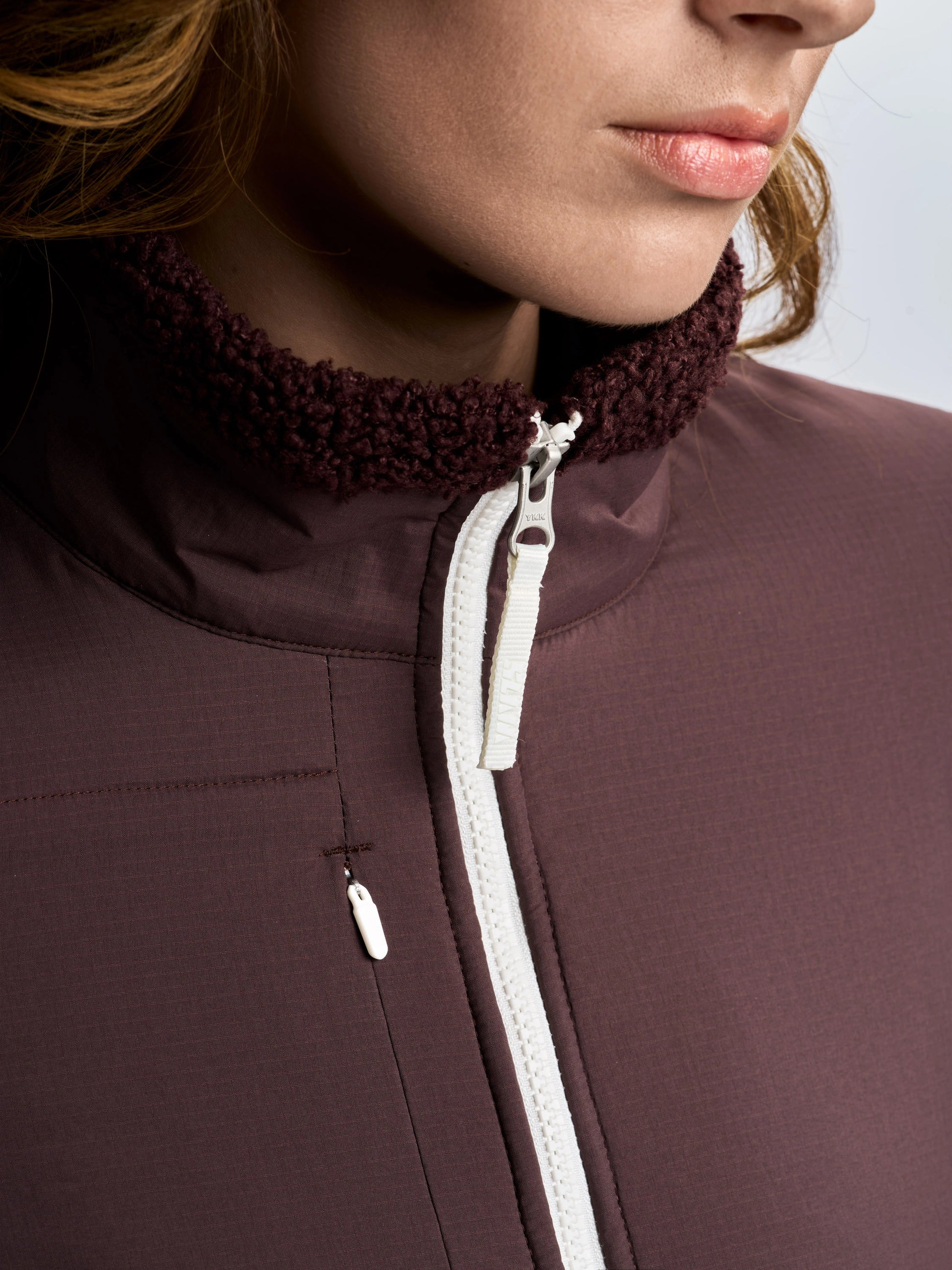 A close-up captures a person wearing the SLAM ALTURA WS FZ FLEECE in maroon, featuring a white zipper and a textured maroon collar that highlights its stylish yet functional design. The vest provides superior wind protection, while the soft fleece lining keeps you warm in cooler conditions. The person’s face is partially visible, with only the lips and chin shown.