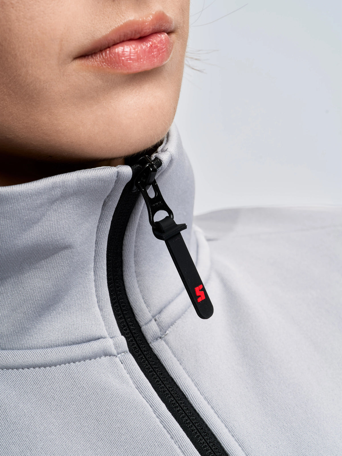 Close-up of a person wearing the SLAM ACT WS FLEECE POWERSTR, a light gray fleece jacket with a black zipper. The zipper pull features a small red “5” detail, while part of the person’s chin and lips are visible—emphasizing the sleek design characteristic of women’s activewear.