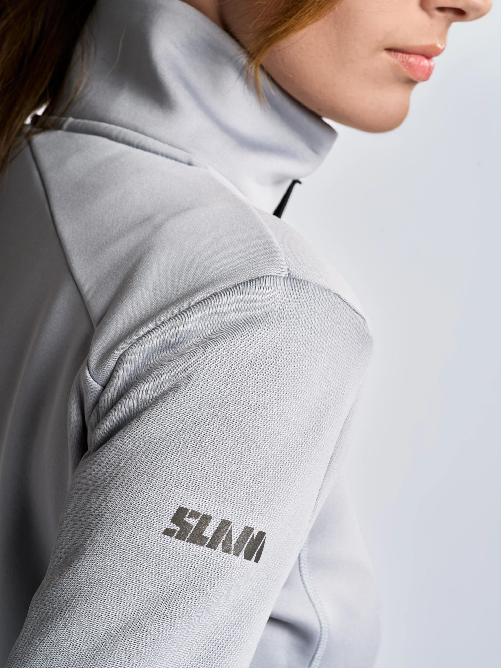 A close-up shows a person wearing the SLAM ACT WS FLEECE POWERSTR jacket in light gray, featuring “SLAM” printed on the left sleeve. Crafted from recycled fabric, this stylish piece elevates women’s activewear. The person’s lower face and part of their hair are visible against a plain background.
