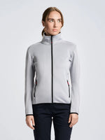 A person stands facing forward, wearing the SLAM ACT WS FLEECE POWERSTR women’s activewear jacket in light gray with black zippers, paired with dark pants. The background is plain and light-colored. The person’s hair is pulled back.