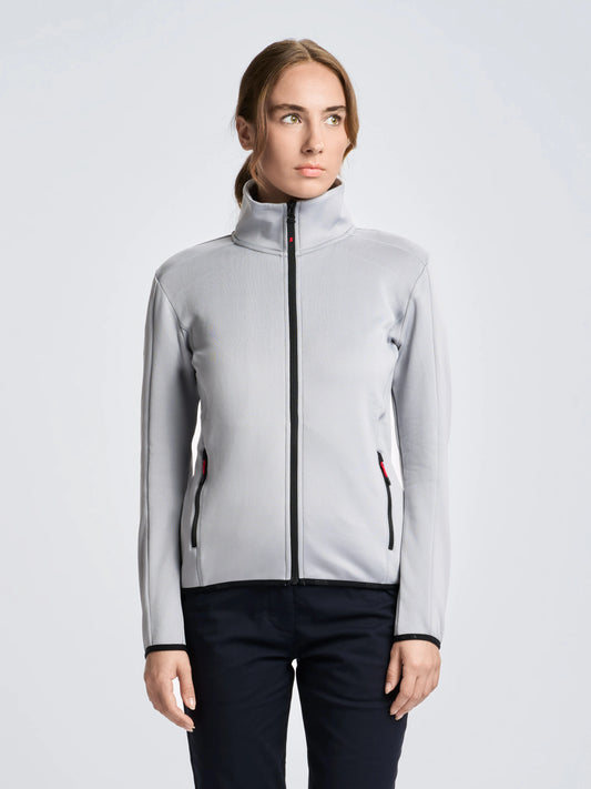 A person stands facing forward, wearing the SLAM ACT WS FLEECE POWERSTR women’s activewear jacket in light gray with black zippers, paired with dark pants. The background is plain and light-colored. The person’s hair is pulled back.