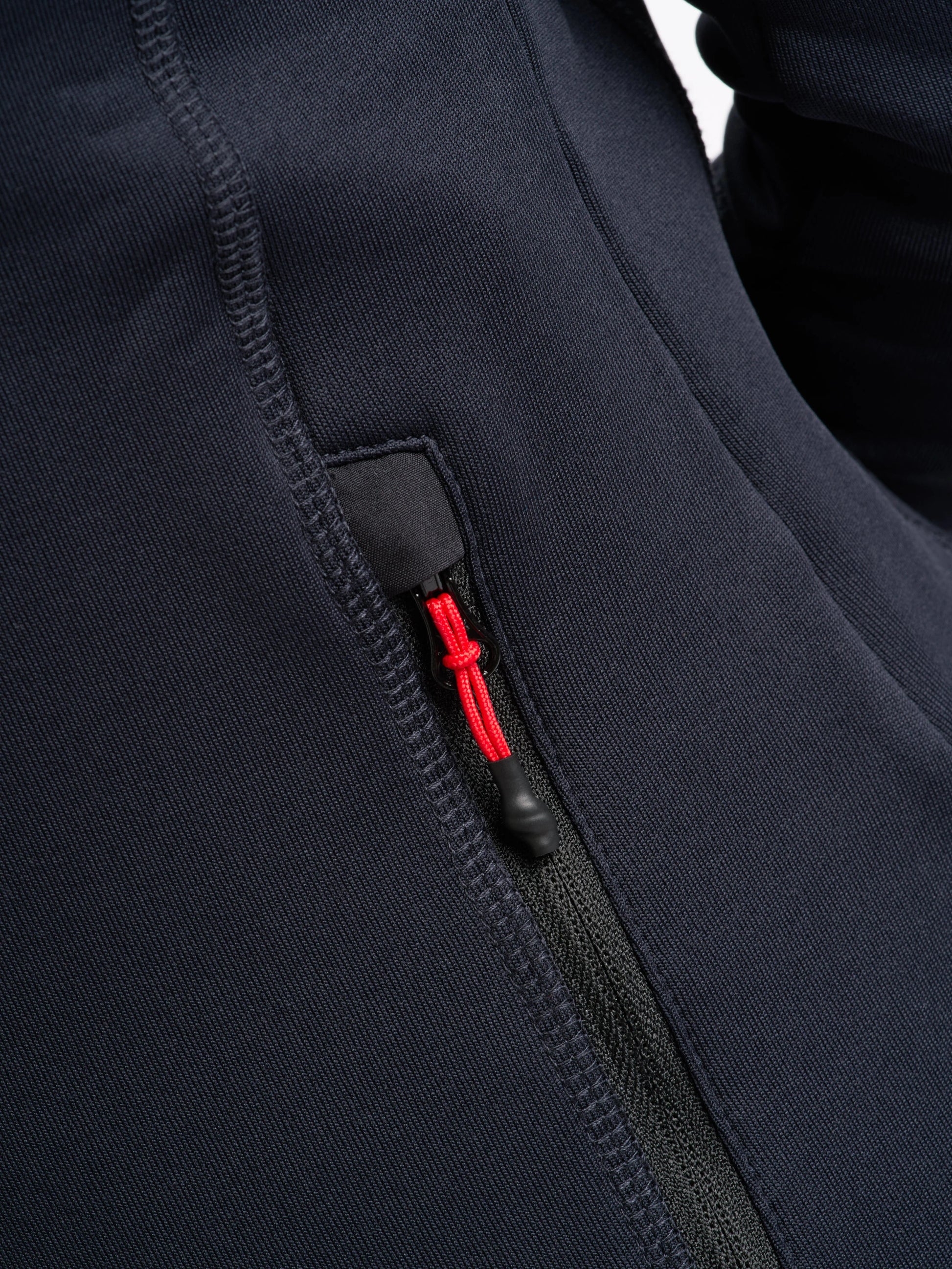 Close-up view of a zippered pocket on the SLAM ACT WS FLEECE POWERSTR in navy blue. The pocket features a black-based zipper with a bright red pull tab, and the garment is crafted from recycled fabric, showcasing visible stitching and a smooth texture.