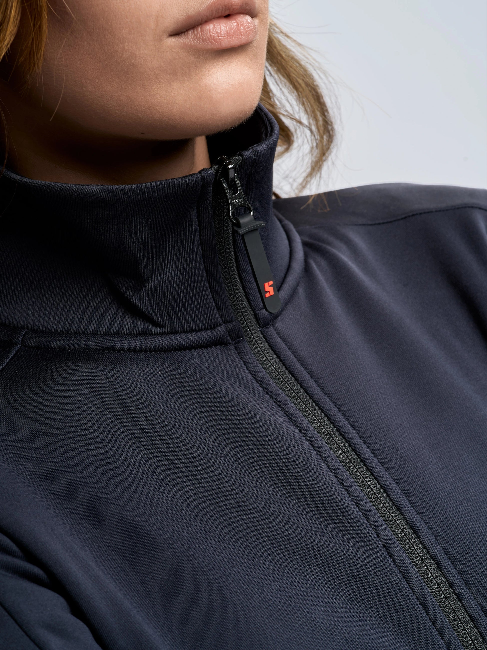 A close-up of a person wearing the SLAM ACT WS FLEECE POWERSTR, a dark technical fleece zip-up jacket with a high collar. The zipper is partially closed and features a small red SLAM logo on the pull tab. Only the lower half of the person’s face is visible.