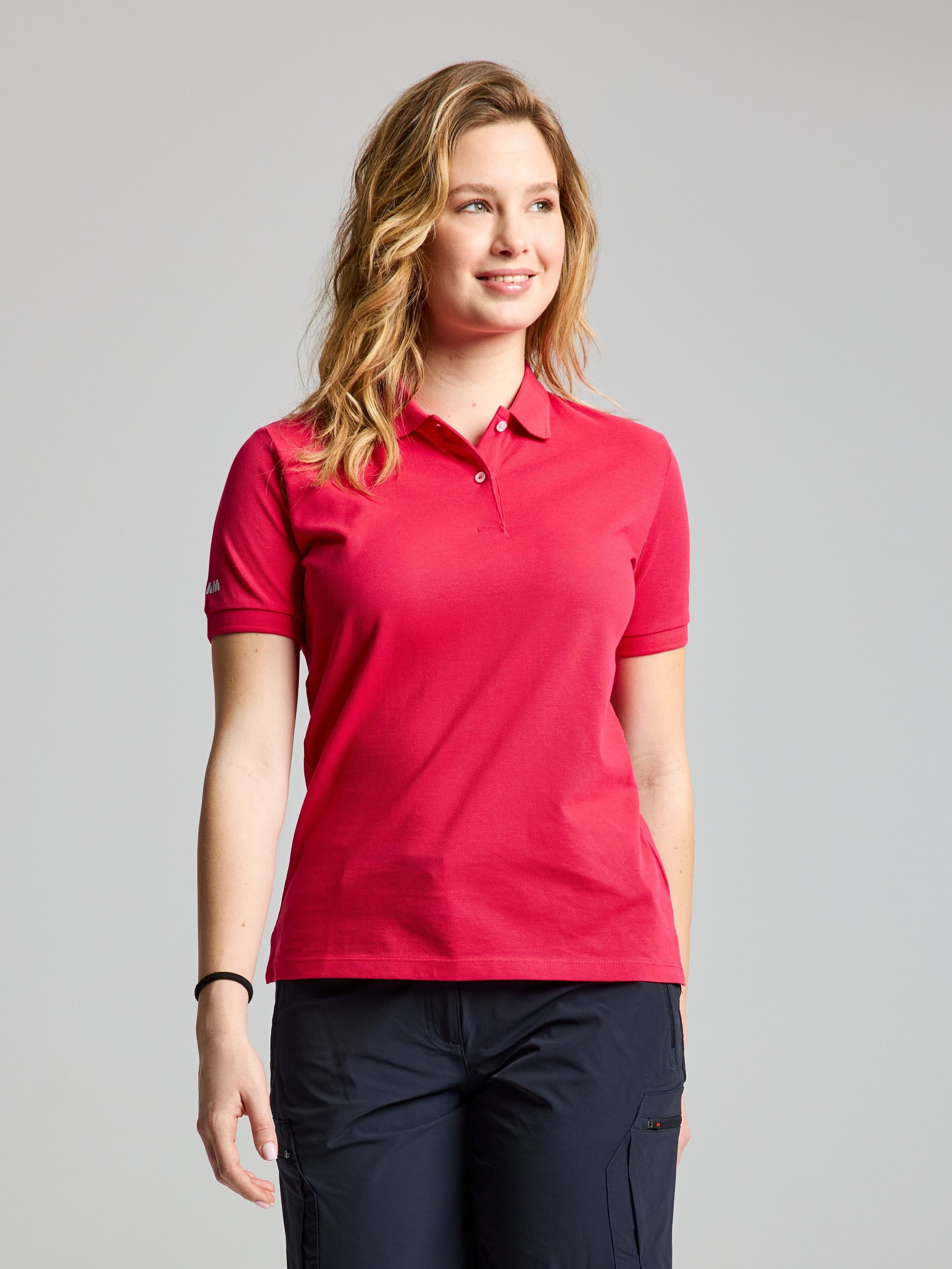 A woman with long hair is wearing a SLAM's DECK WOMAN POLO SHORT SLEEVE in VIRTUAL PINK paired with dark pants. She looks to her left, set against a plain light background, highlighting her stylish outfit.