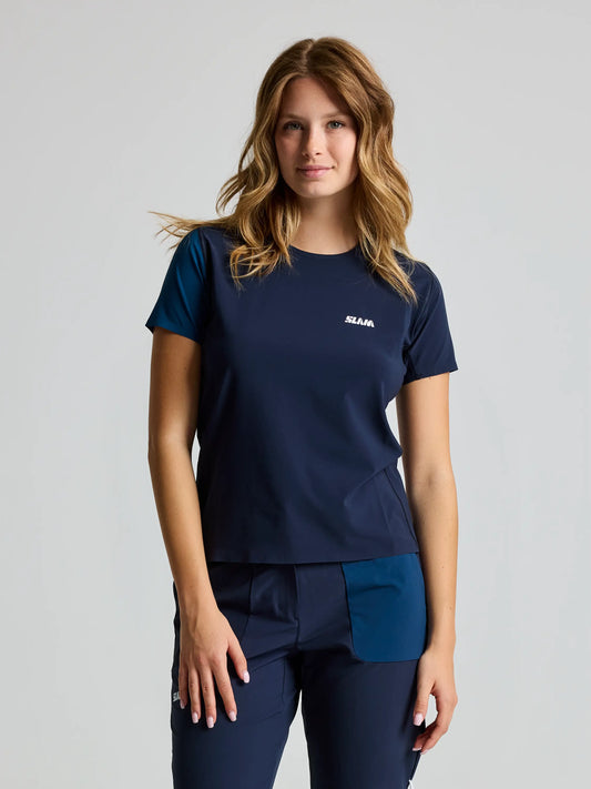 Against a plain background, a person with shoulder-length wavy hair wears an OD WS TECH T-SHIRT SS by SLAM, featuring UV 50+ protection and a small white logo on the chest. They are in navy blue short sleeves and pants, looking directly at the camera.