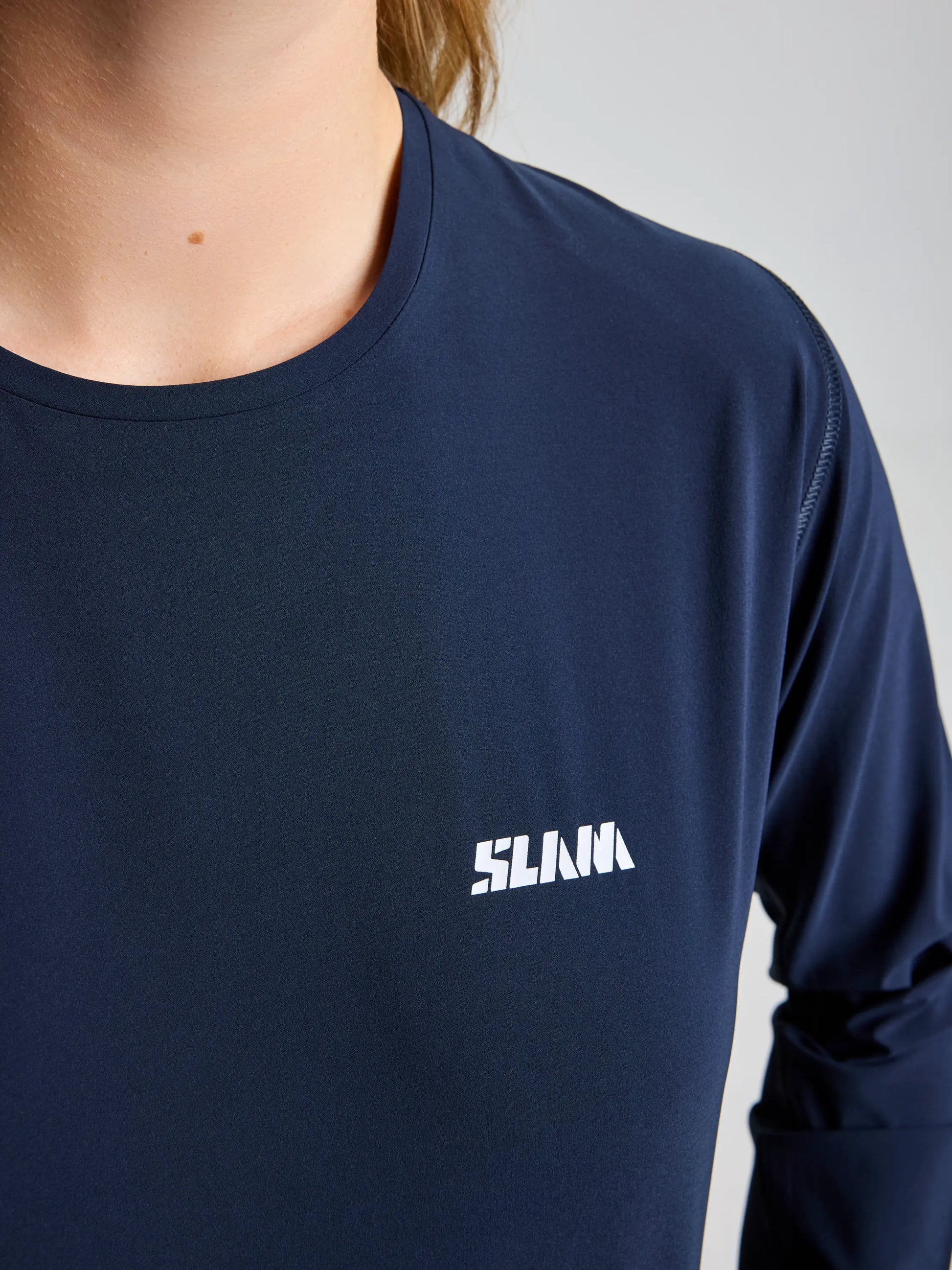 A person wearing the SLAM OD WS TECH T-SHIRT LS in navy blue with "SLAM" printed in white on the left chest. Only the lower face, neck, and upper torso are visible.