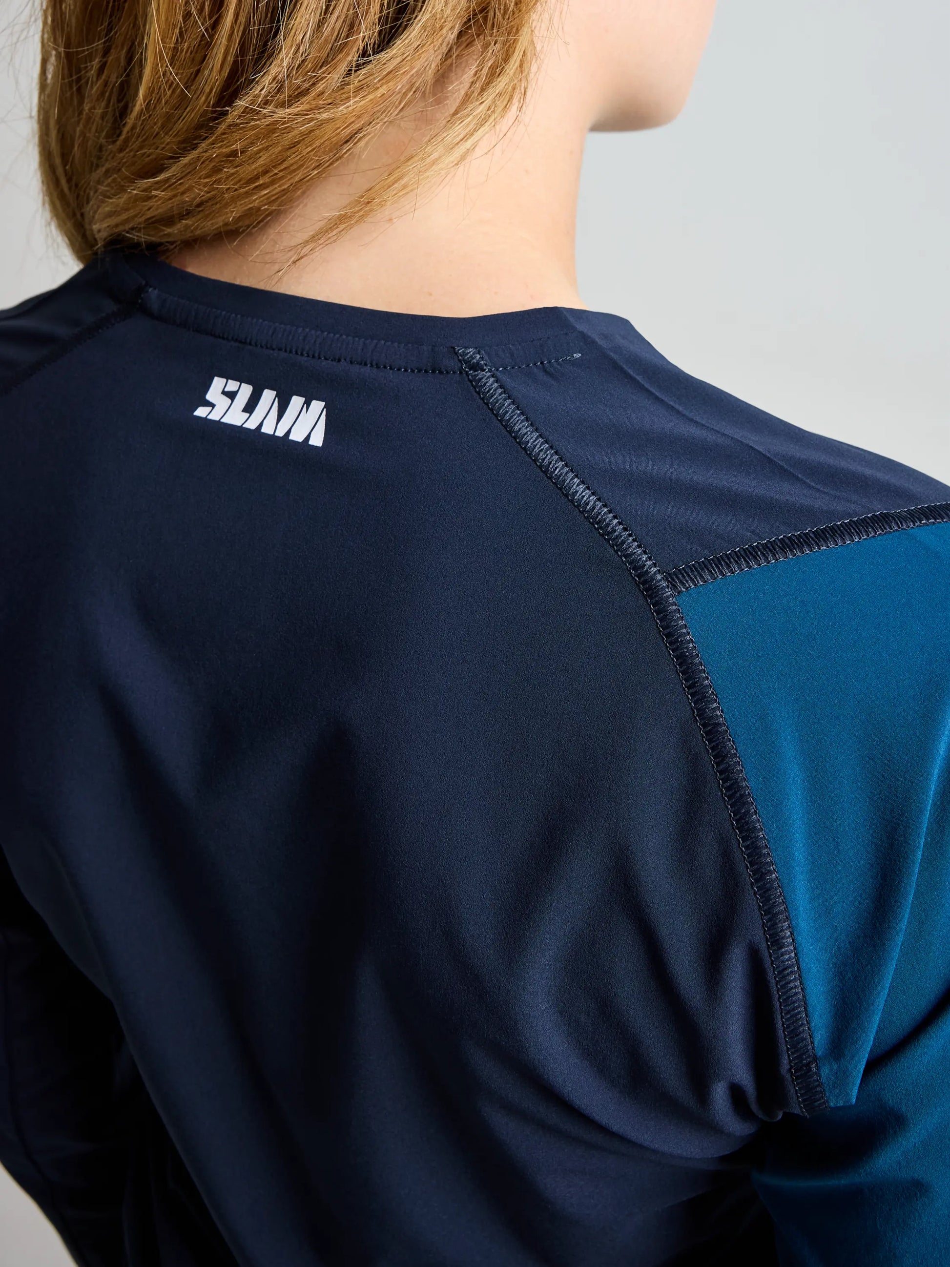 A person with light brown hair is seen from behind wearing the SLAM OD WS TECH T-SHIRT LS in navy blue and teal, featuring "SLAM" printed in white near the neckline on the back. This shirt offers quick-dry performance and modern style.