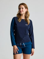 A woman wears the SLAM OD WS TECH T-SHIRT LS, a navy blue long-sleeve swim shirt with UV protection, and matching shorts. She stands against a plain light background, looking slightly to the side with a neutral expression.