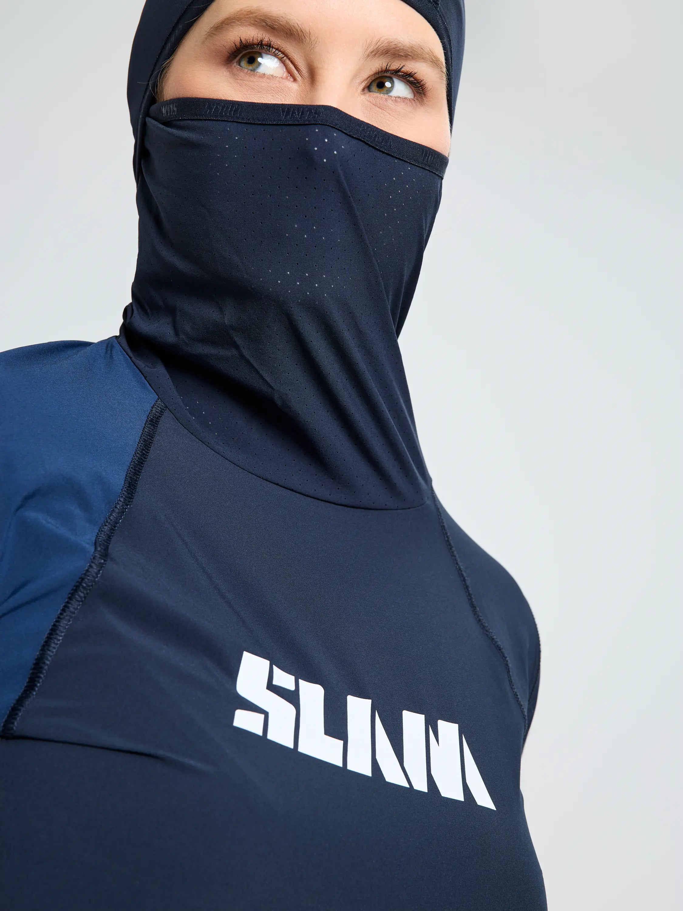 A person wears the SLAM OD WS TECH HOODED T-SHIRT, a navy blue sports hijab and face covering made from recycled stretch fabric, with "SLAM" in white on the front, set against a plain light-colored background.