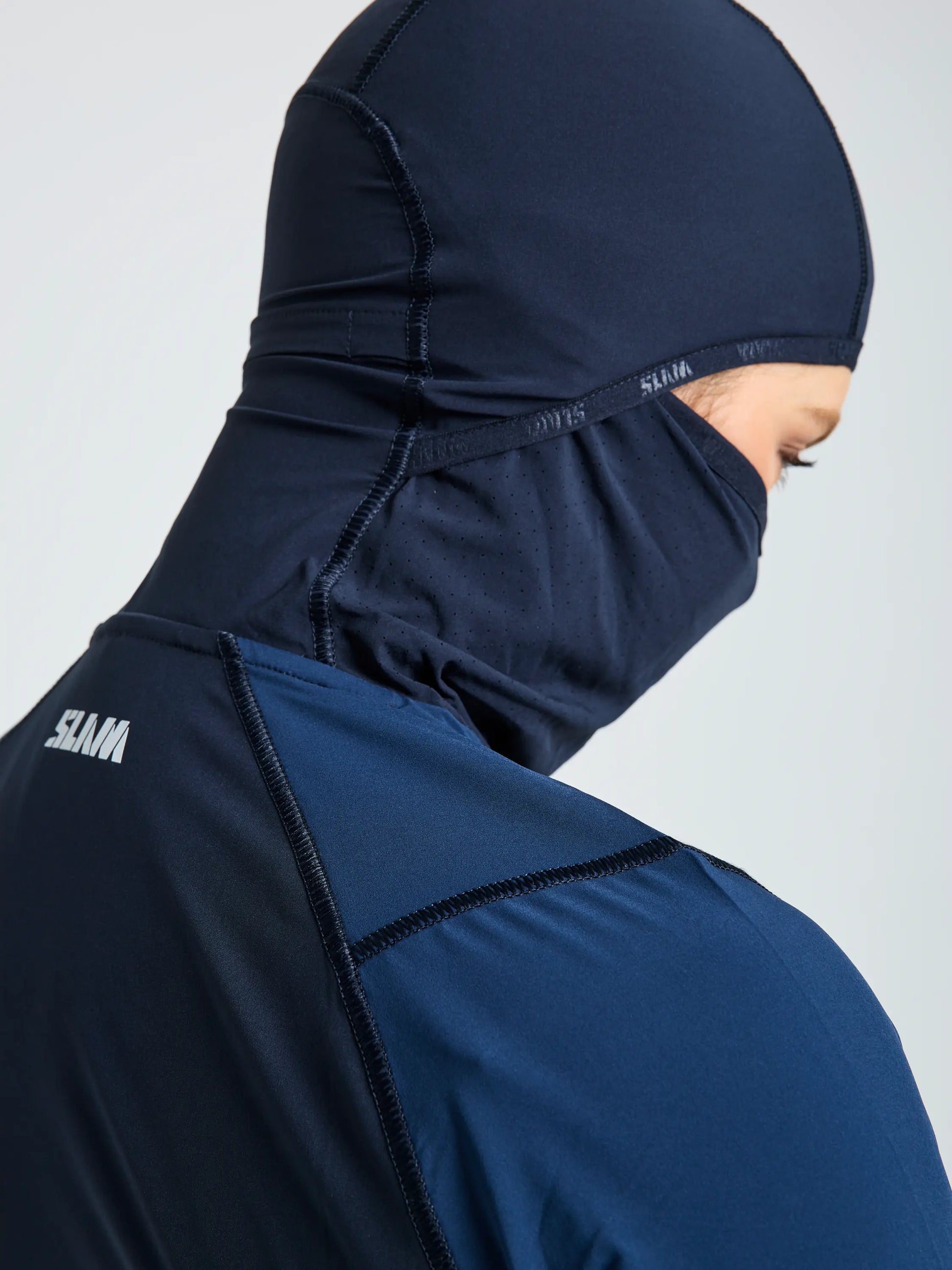 A person wears the SLAM OD WS TECH HOODED T-SHIRT in navy, covering head, neck, and lower face. "SLAM" is printed in white on the back. Made from recycled stretch fabric. The background is plain and light-colored.