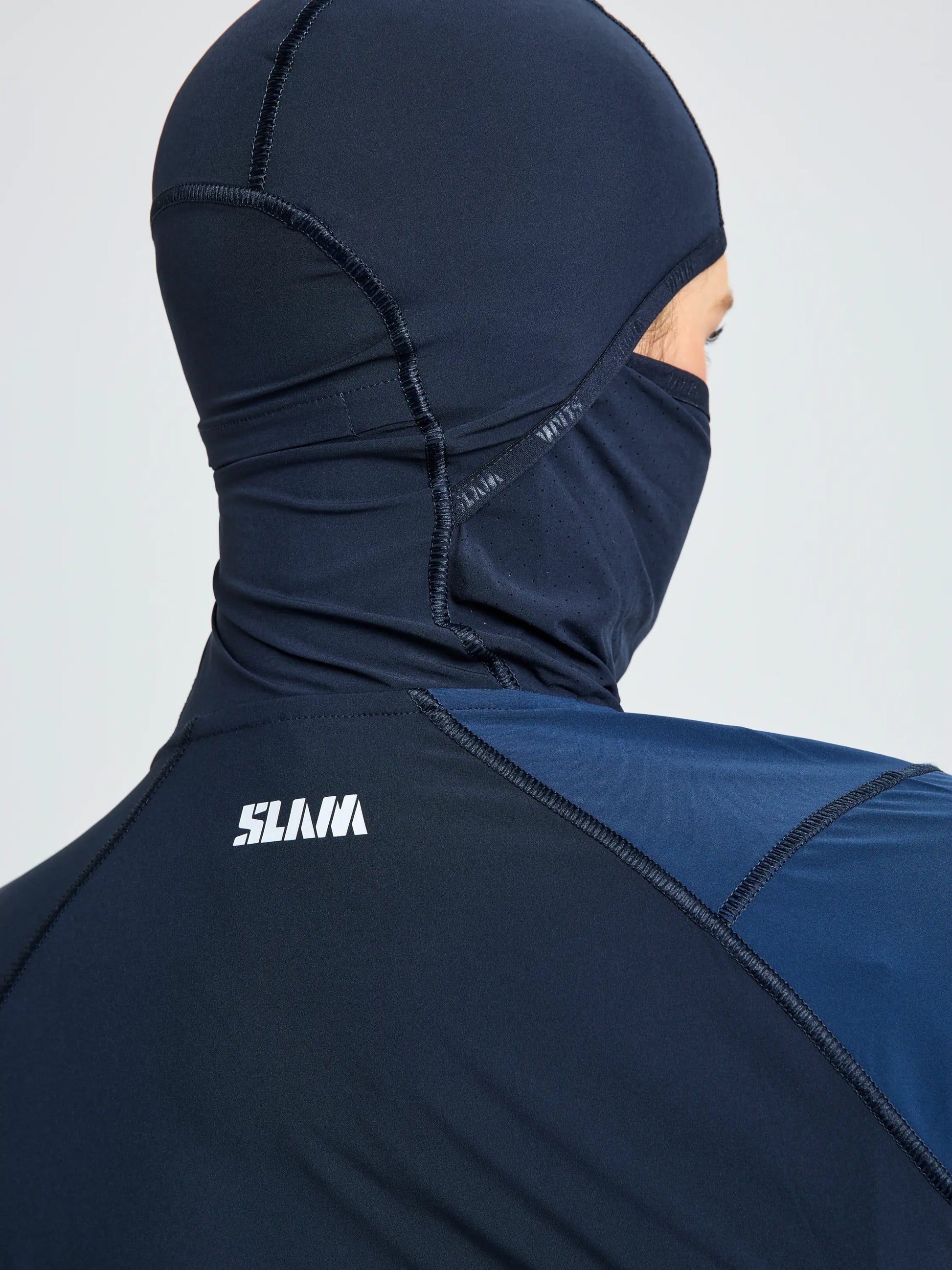 A person is seen from the back wearing the SLAM OD WS TECH HOODED T-SHIRT in navy blue, made from recycled stretch fabric with a face covering. "SLAM" is printed in white on the upper back of the garment.