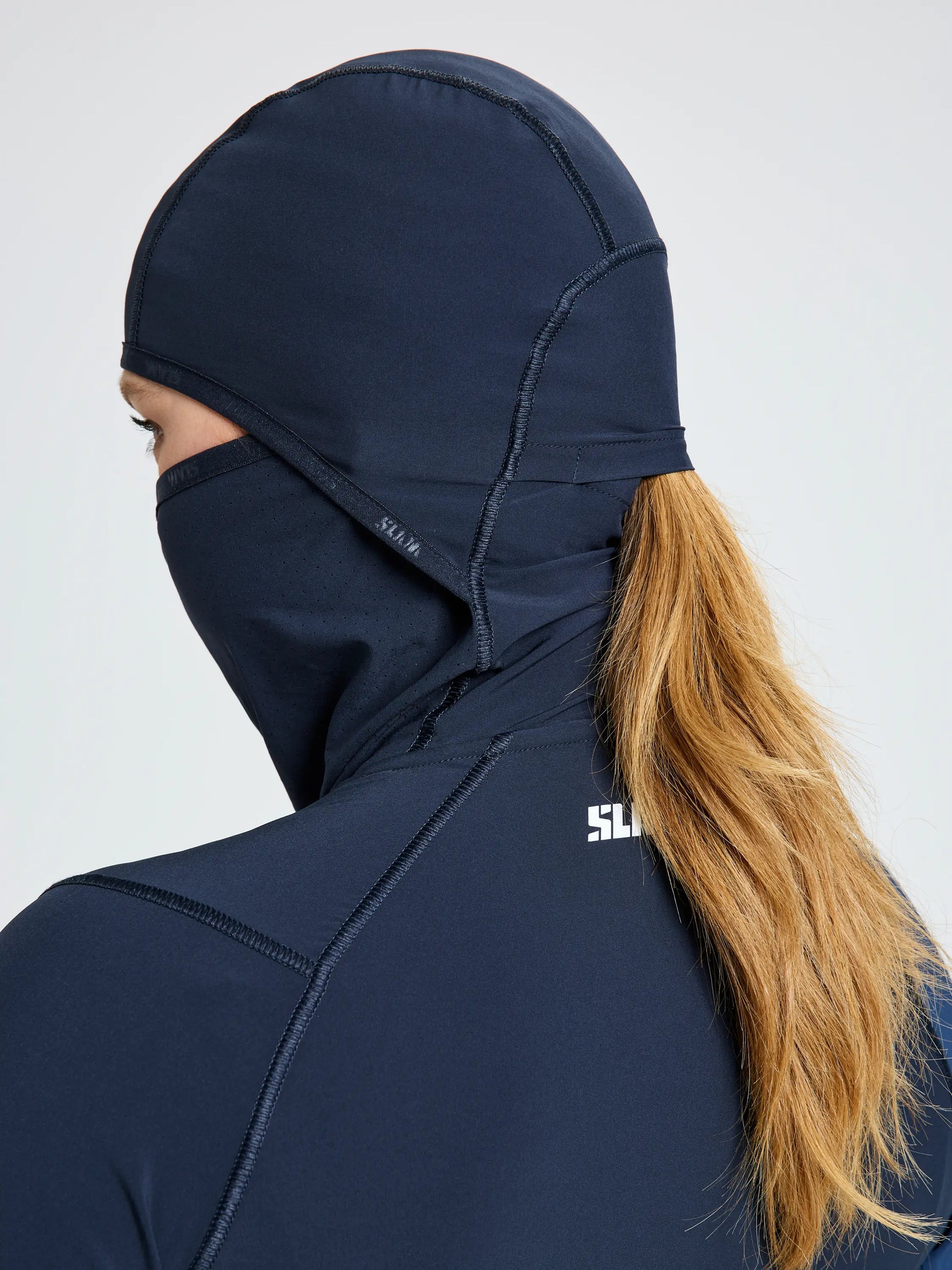 A person with long blond hair wears the SLAM OD WS TECH HOODED T-SHIRT in navy blue, which covers their head, neck, and lower face. The shirt is made from recycled stretch athletic fabric and leaves only part of the face exposed.