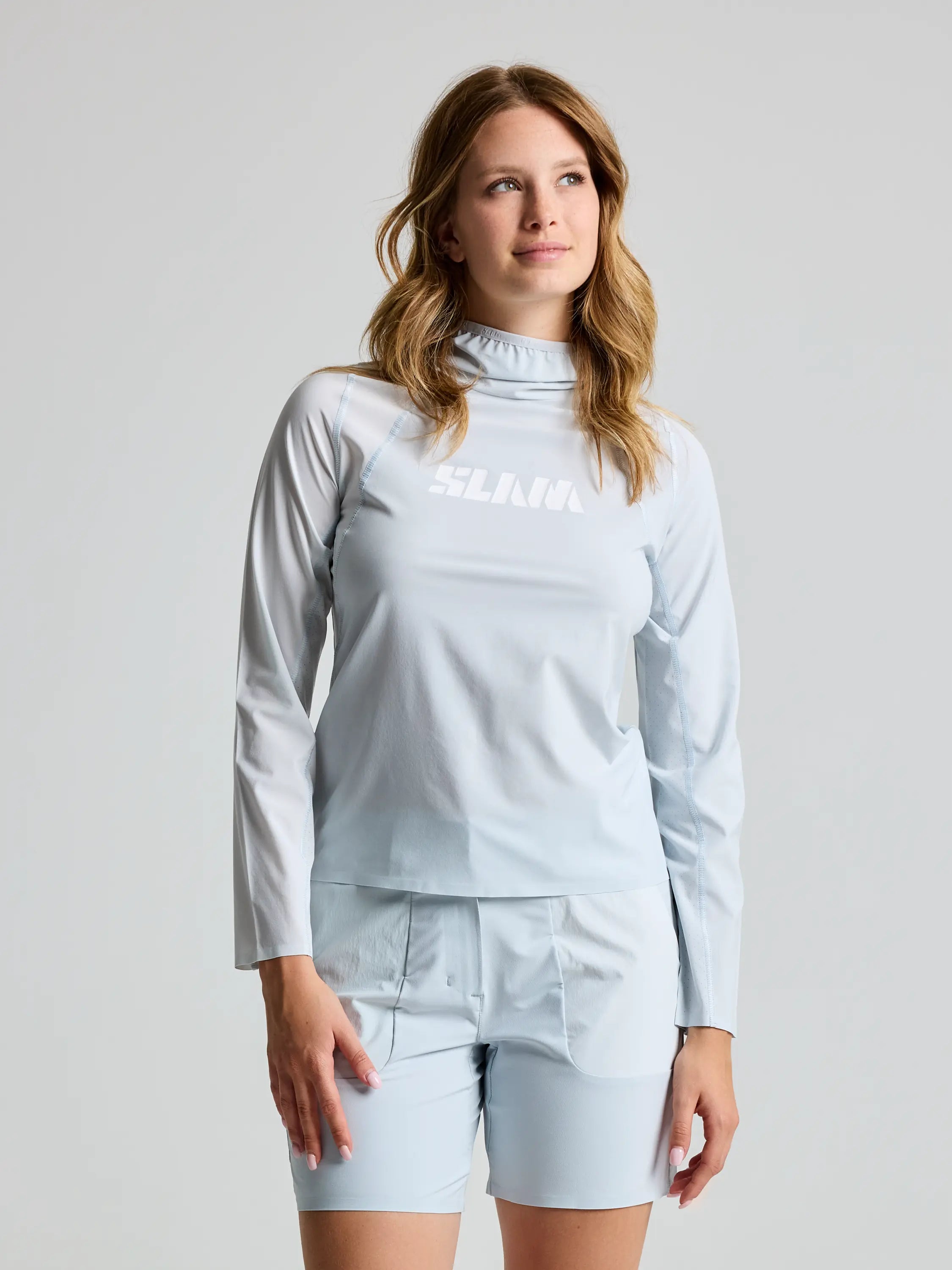 A woman stands wearing a CLIFF GREY OD WS TECH HOODED T-SHIRT and matching shorts by SLAM, made from recycled stretch fabric. A white logo adorns the chest of the top, which complements her wavy hair as she gazes off to the side against a plain background.