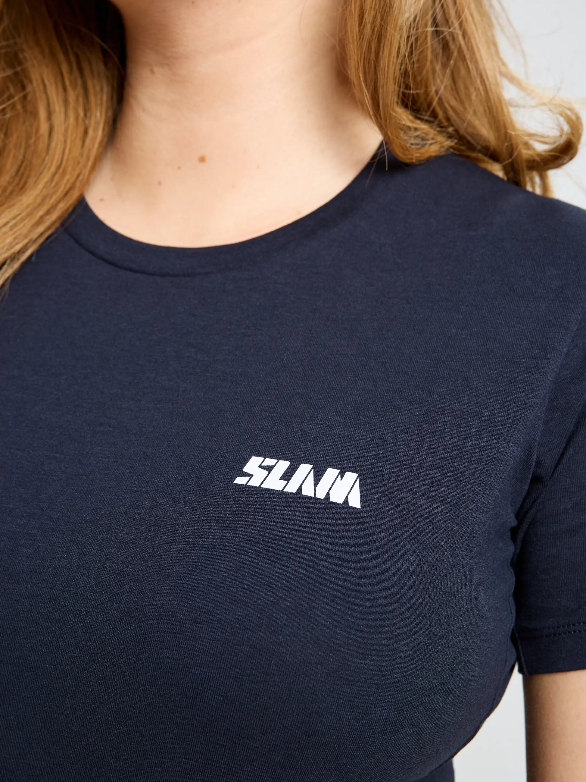 A person with long light brown hair wears the SLAM GRAPHIC WS T-SHIRT SS in dark blue bamboo cotton, featuring "SLAM" in bold white letters on the left chest. The image is cropped to show only the upper torso and neckline.